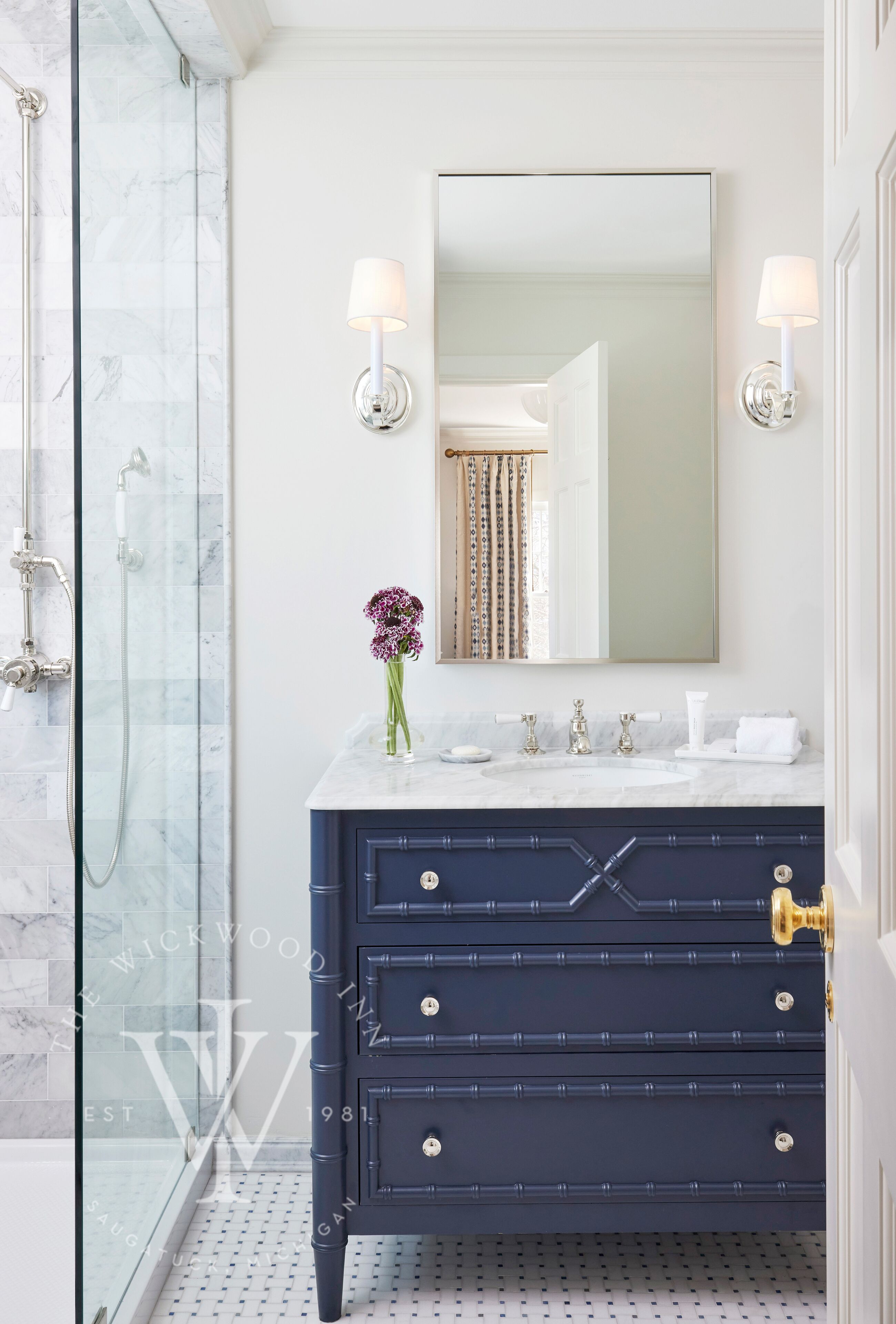 A bathroom with a glass shower enclosure, a marble-tiled wall, a dark blue vanity with gold handles, a white sink, and a mirror with wall sconces.