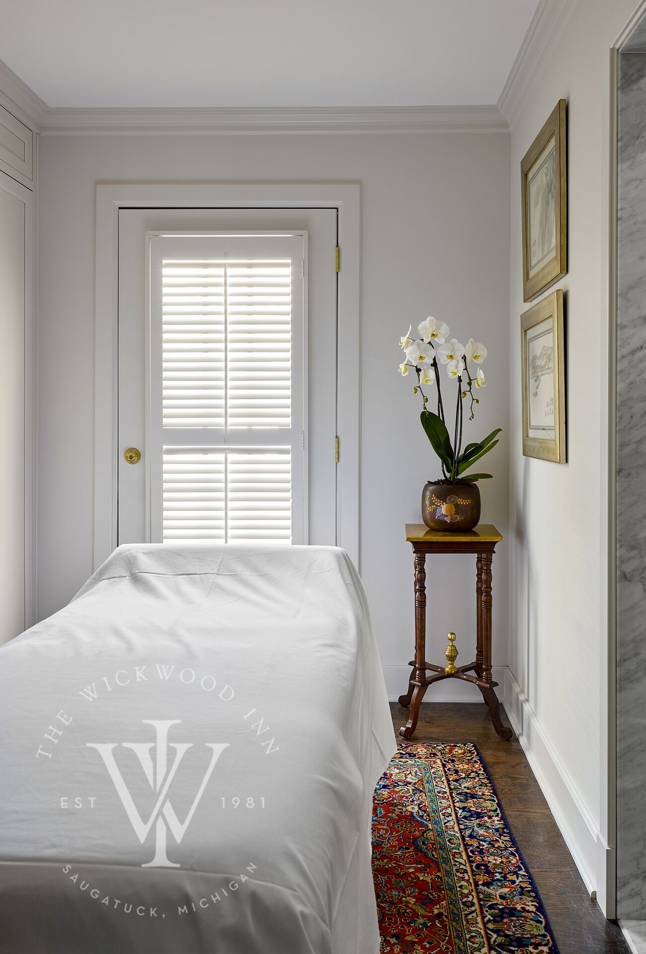 A hotel room with a neatly made bed, a small side table with a vase of flowers, a patterned rug, and a door with blinds.