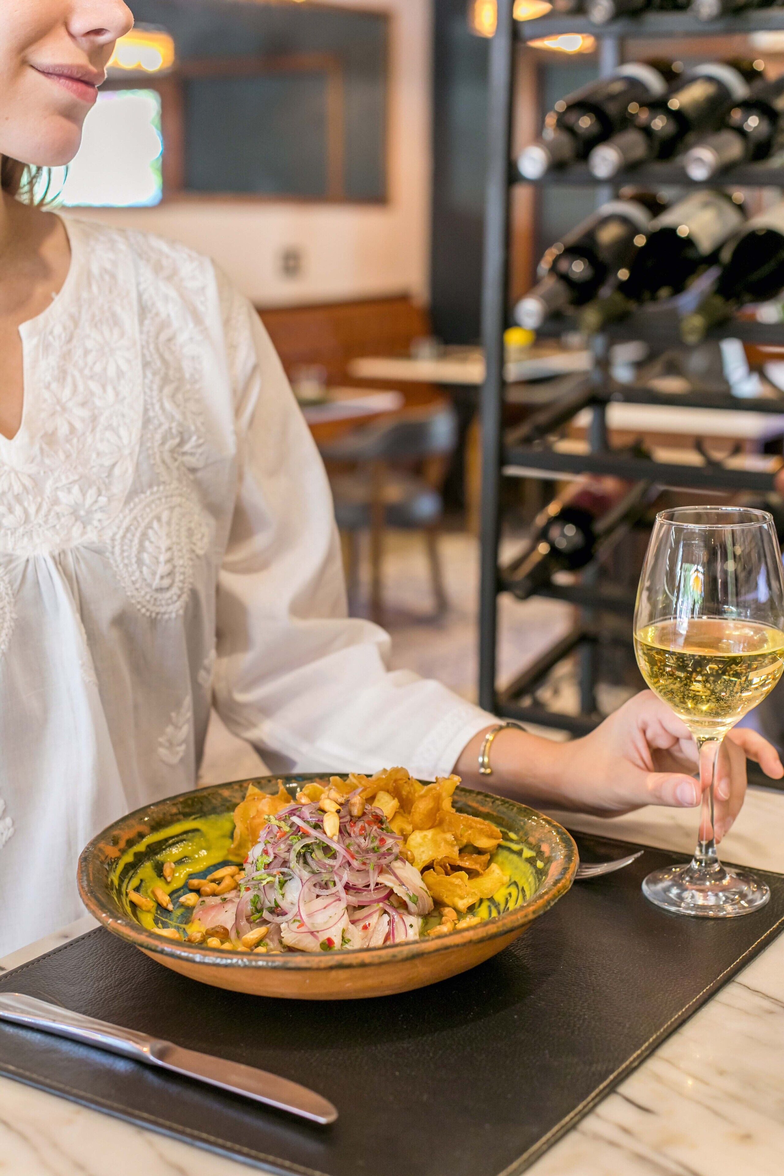 Une personne tenant un verre de vin blanc à côté d’un bol de nourriture, avec un couteau et une cuillère posés sur la table.