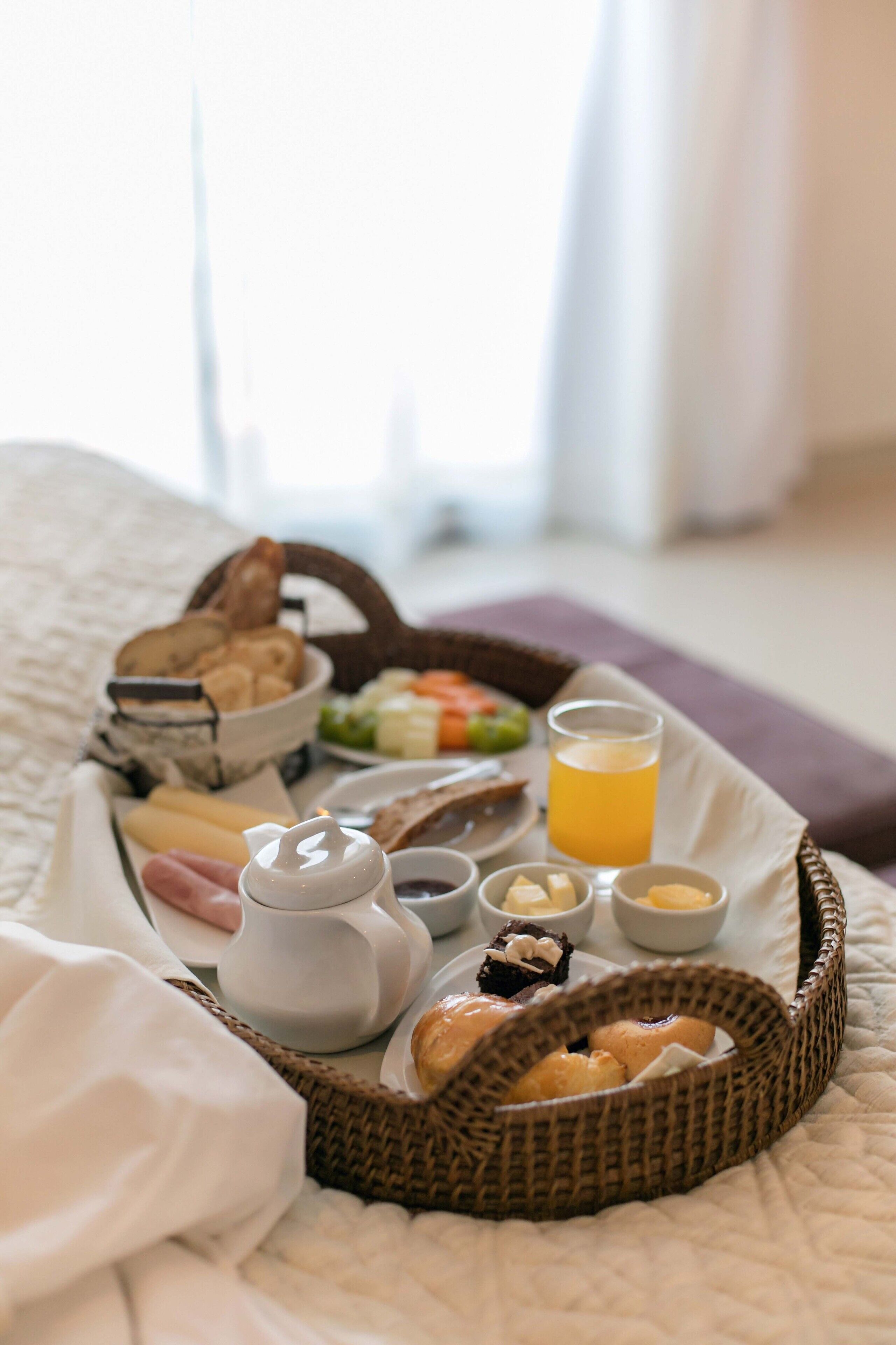 Un plateau de petit-déjeuner sur un lit, avec du pain, des viennoiseries, du fromage et un verre de jus d’orange.