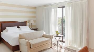 A hotel room with a large bed, a sofa, a small table with a plant, and a view of greenery through the window.