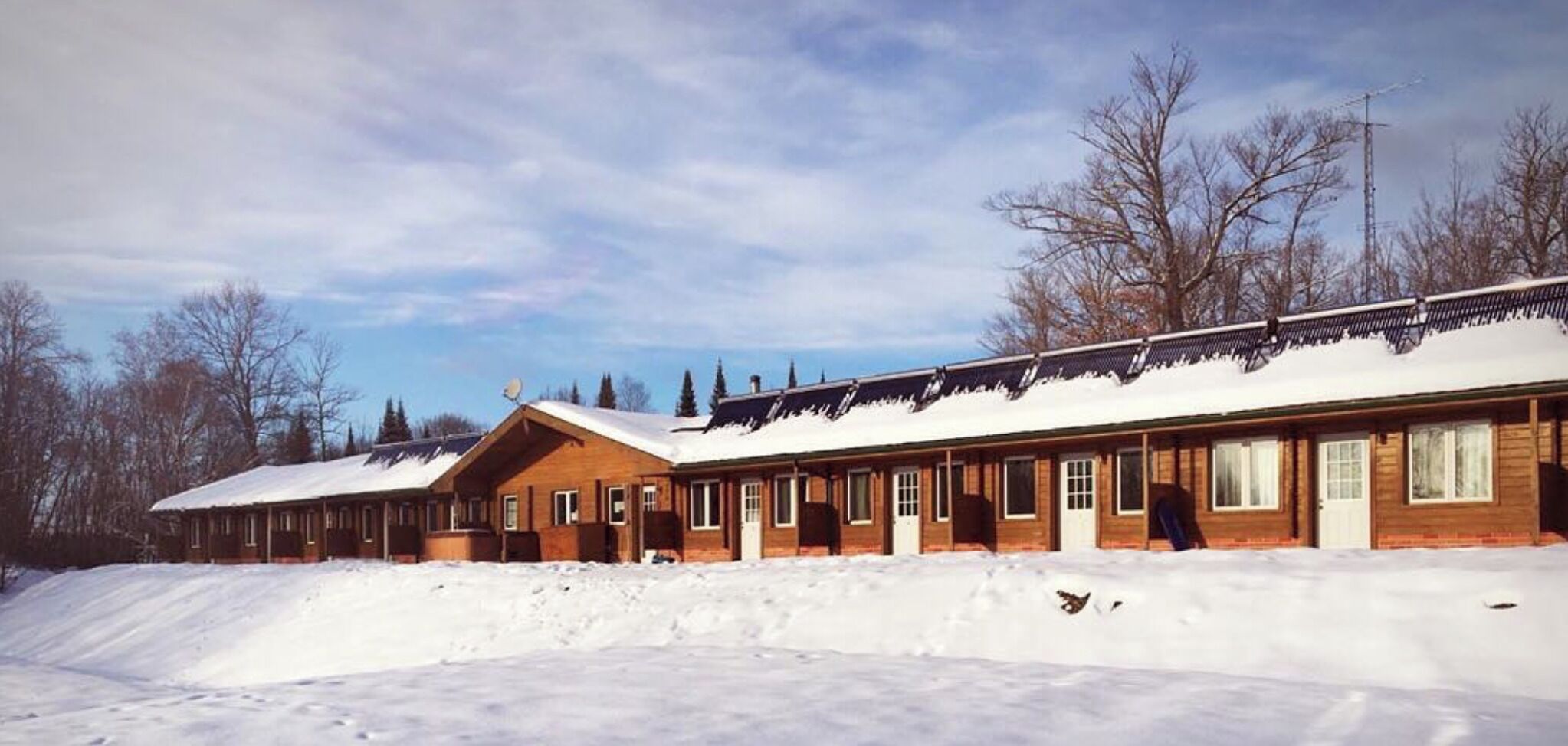 Une rangée de chalets en bois aux toits enneigés, nichés dans un paysage hivernal où les arbres sont dénudés et le ciel est dégagé.
