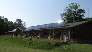 A building with a long roof equipped with solar panels, a small porch area with chairs, and a large tree in the background. - Quiet Bay: Lakeside Escape (Magnetawan)
