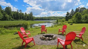 A lakeside outdoor seating area with red Adirondack chairs arranged around a stone fire pit. - Quiet Bay: Lakeside Escape (Magnetawan)