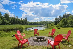 Un espace extérieur aménagé au bord du lac, avec des chaises Adirondack rouges disposées autour d’un foyer en pierre.