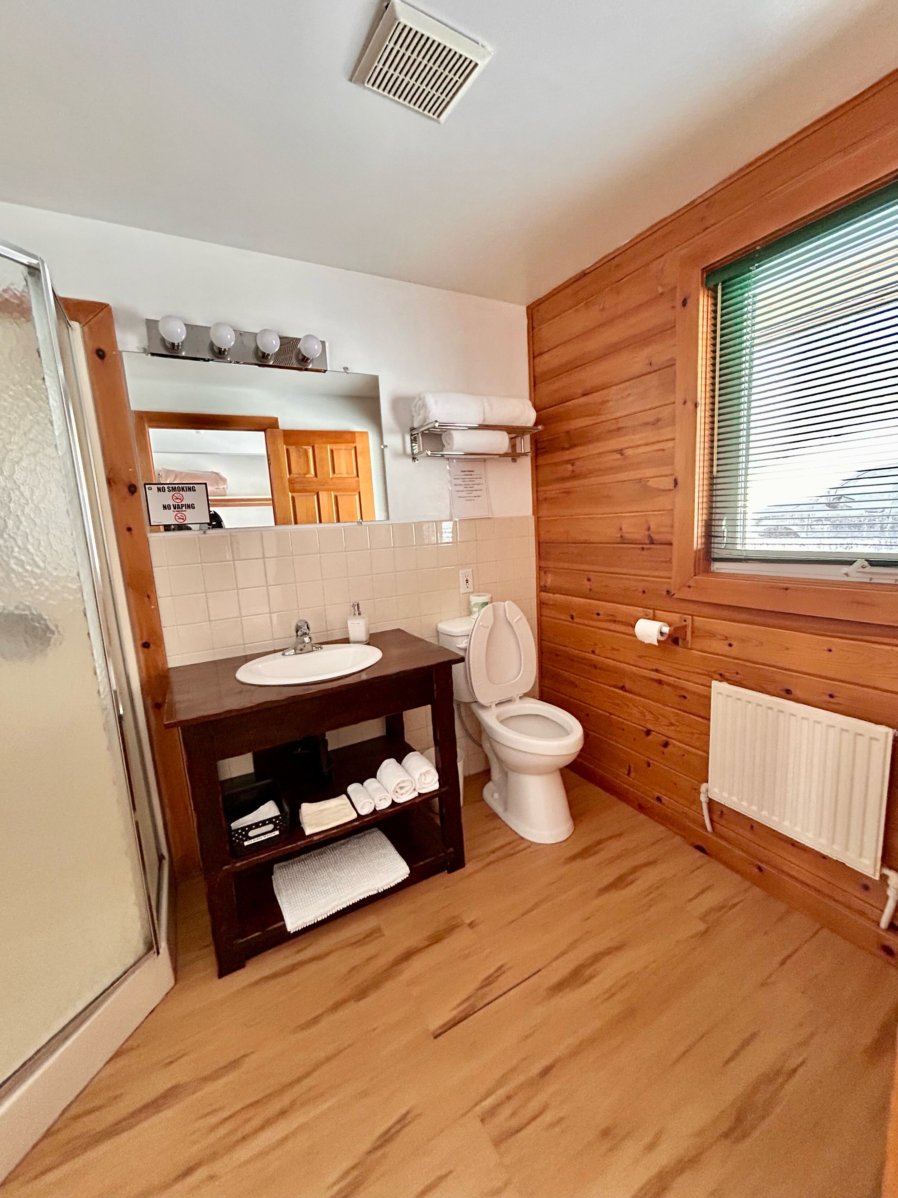 Une salle de bain avec un plancher en bois, un lavabo blanc et des toilettes. On y trouve un porte-serviettes et un miroir au-dessus du lavabo.