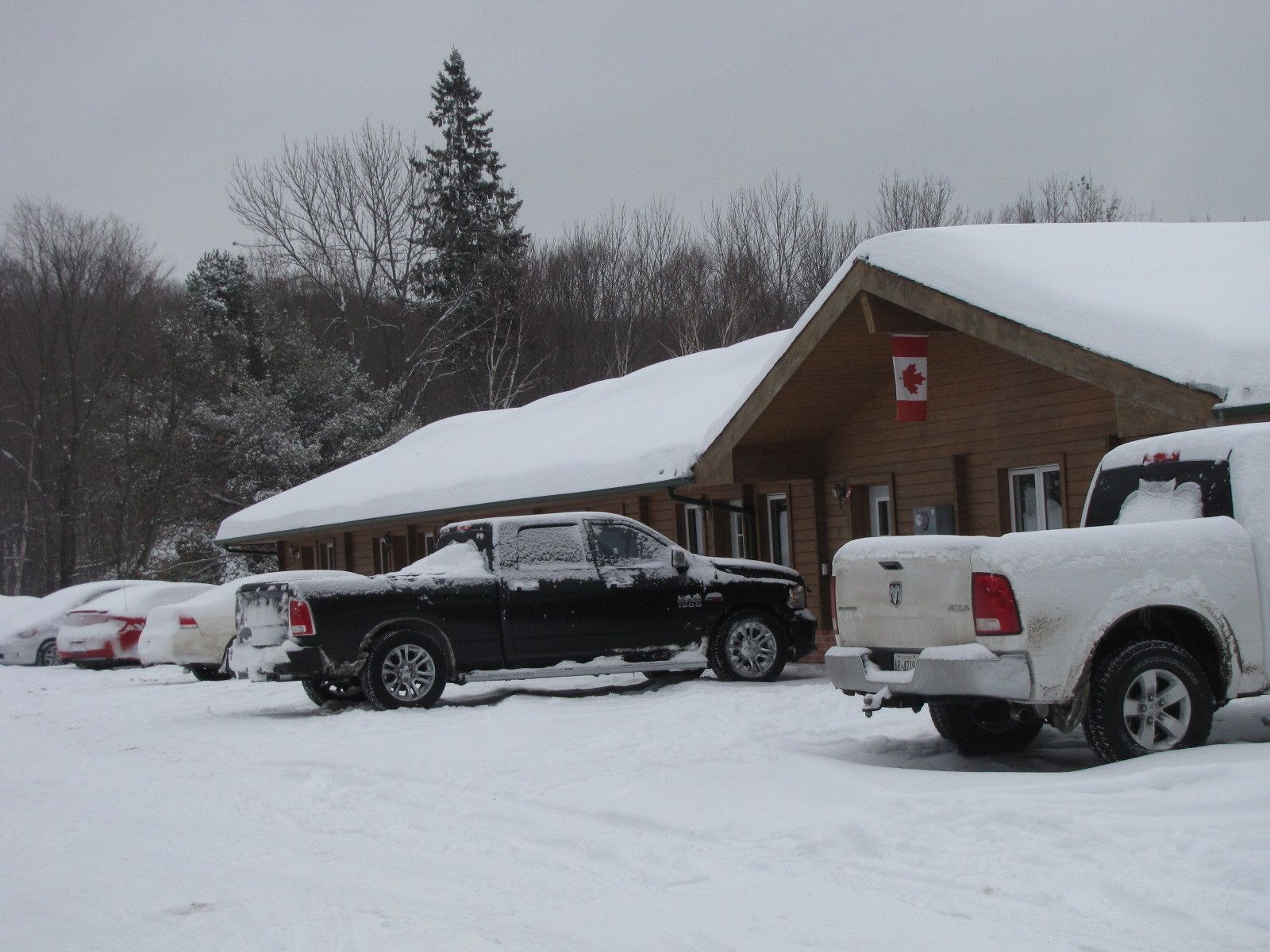 Una cabaña con techo nevado y una bandera canadiense, rodeada de vehículos estacionados.