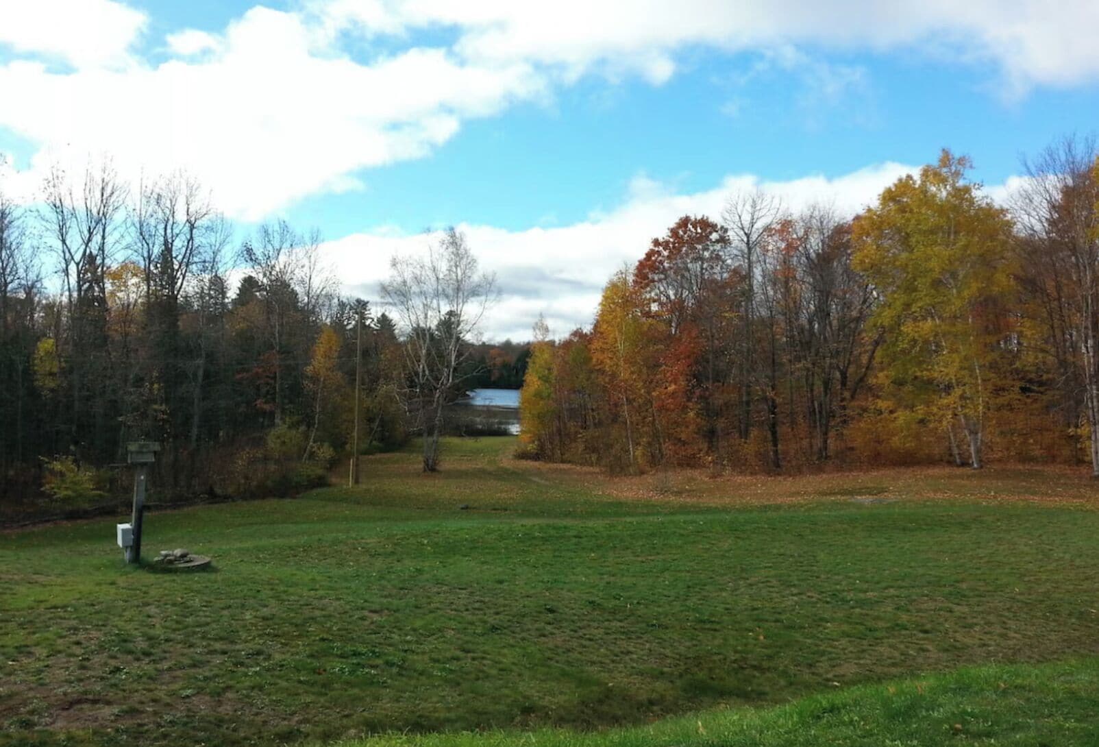 Ein Wiesenfeld mit Bäumen in Herbstfarben, einem Wegweiser und einem Gewässer in der Ferne.