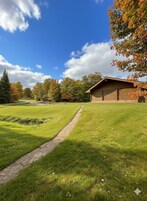 Un sentier bordé d’herbe mène à un bâtiment en bois doté d’un toit en pente, entouré d’arbres aux feuilles d’automne.