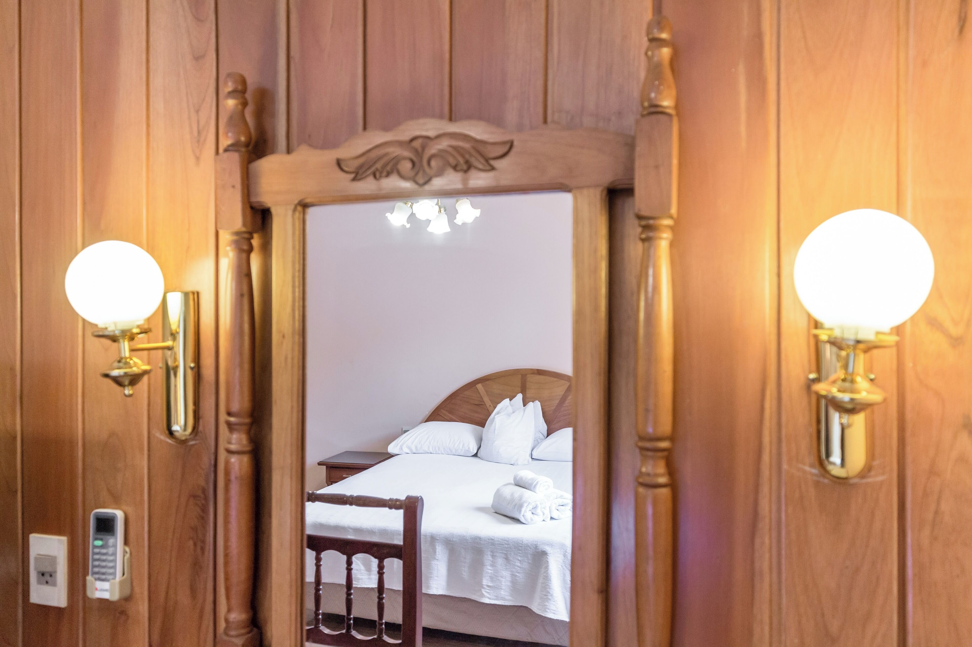 Un marco de madera enmarca un acceso a una cama con sábanas blancas, una mesita de noche y lámparas fijadas a la pared.