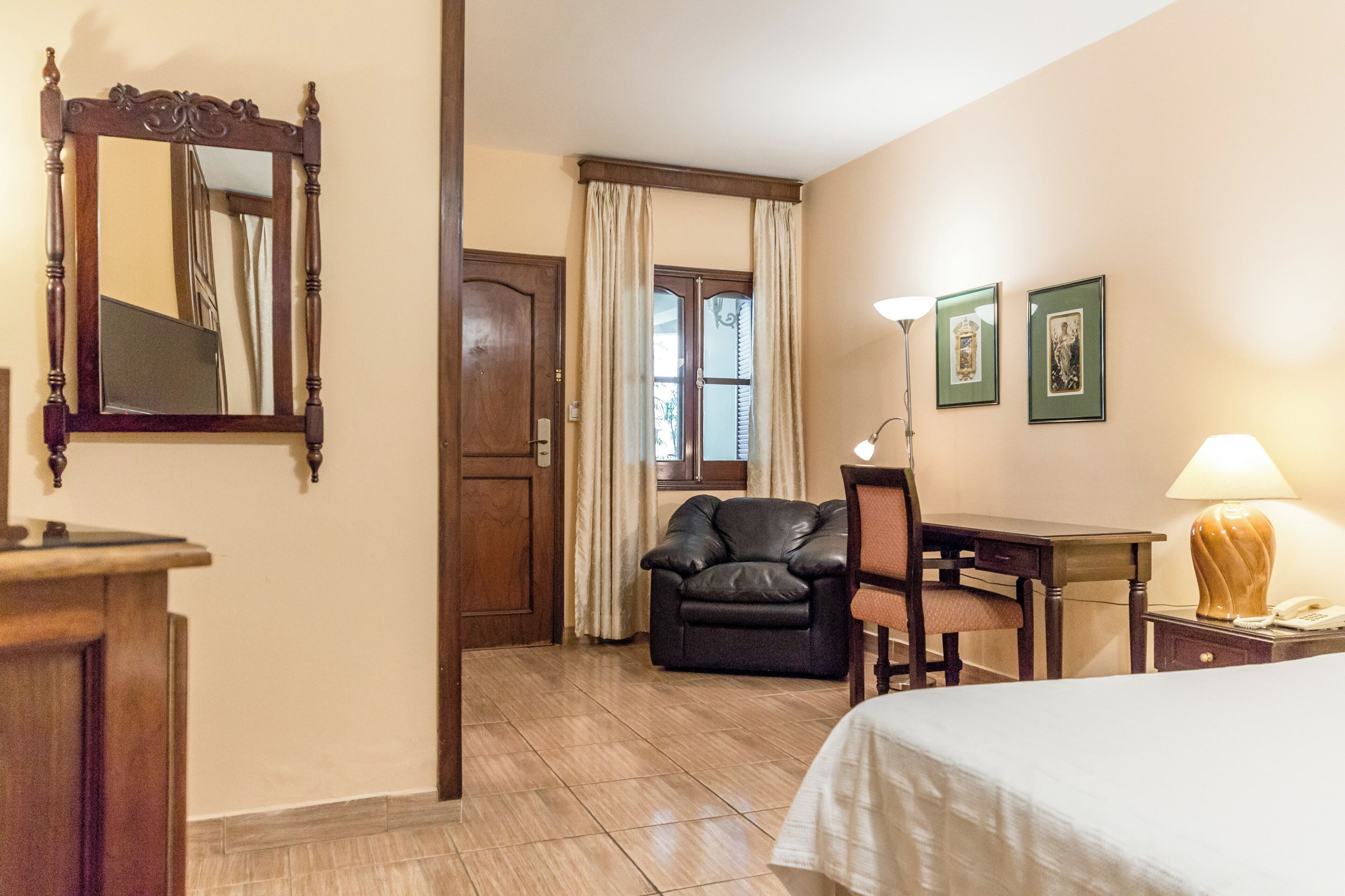 Habitación de hotel con cama, escritorio, silla, sofá de cuero, puerta de madera, espejo y ventana con cortinas.