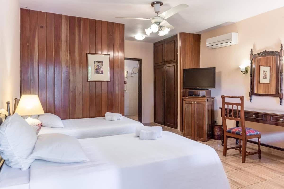 A hotel room with two beds, a desk with a chair, a television, and a ceiling fan.
