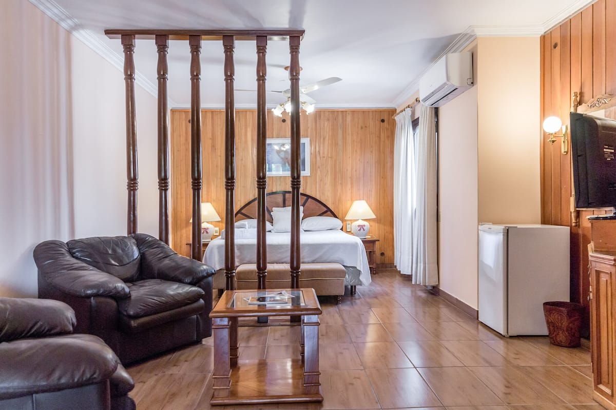 A hotel room with a wooden floor, a bed with a wooden headboard, a leather sofa, a small table, a refrigerator, and a television.