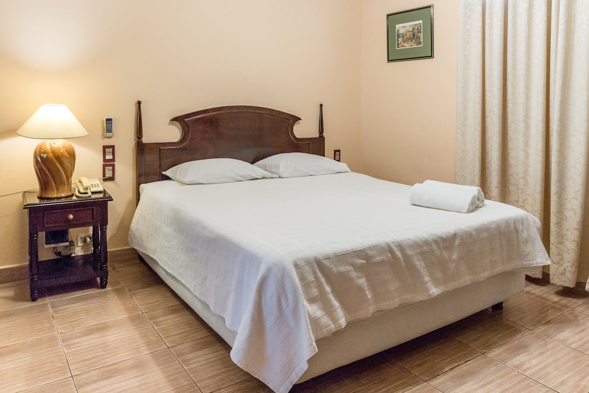 A neatly made bed with a white comforter and pillows, a bedside lamp, a telephone, and a framed picture on the wall.