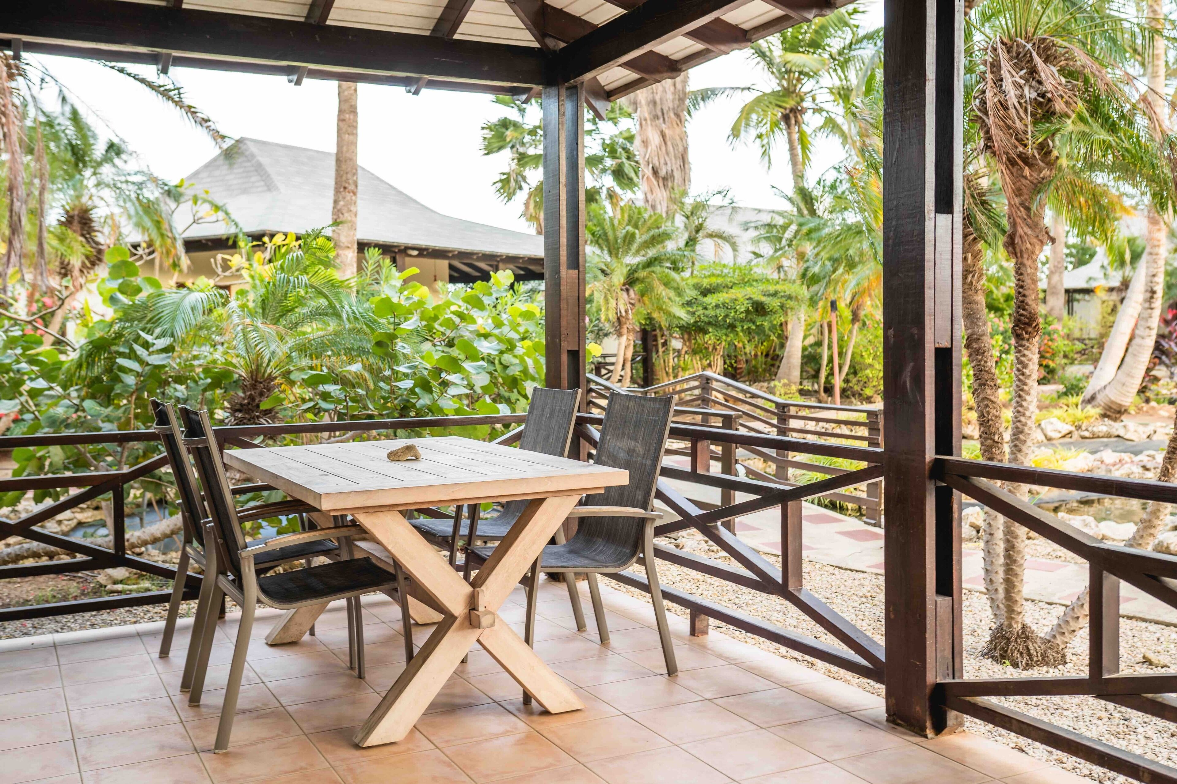 An outdoor patio with a wooden table and chairs, surrounded by a wooden fence and palm trees.