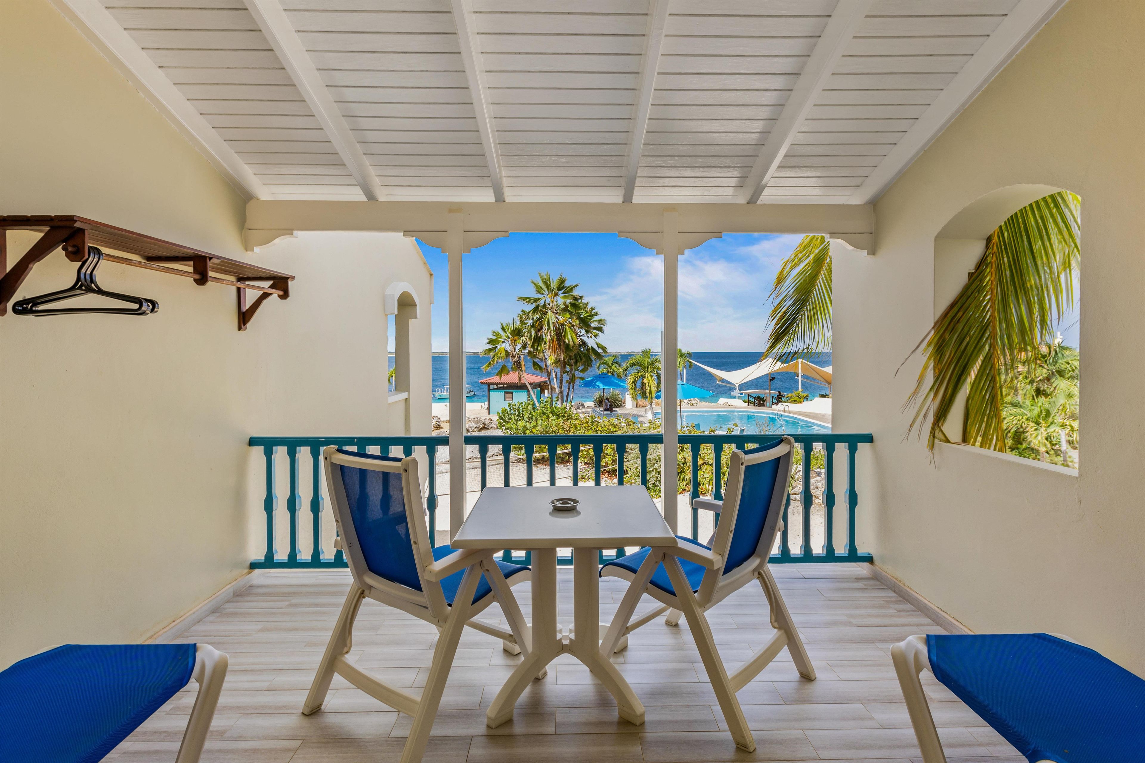 A room with a view of a beach and pool, featuring a table and chairs, a hanging coat rack, and a window with a view of palm trees.