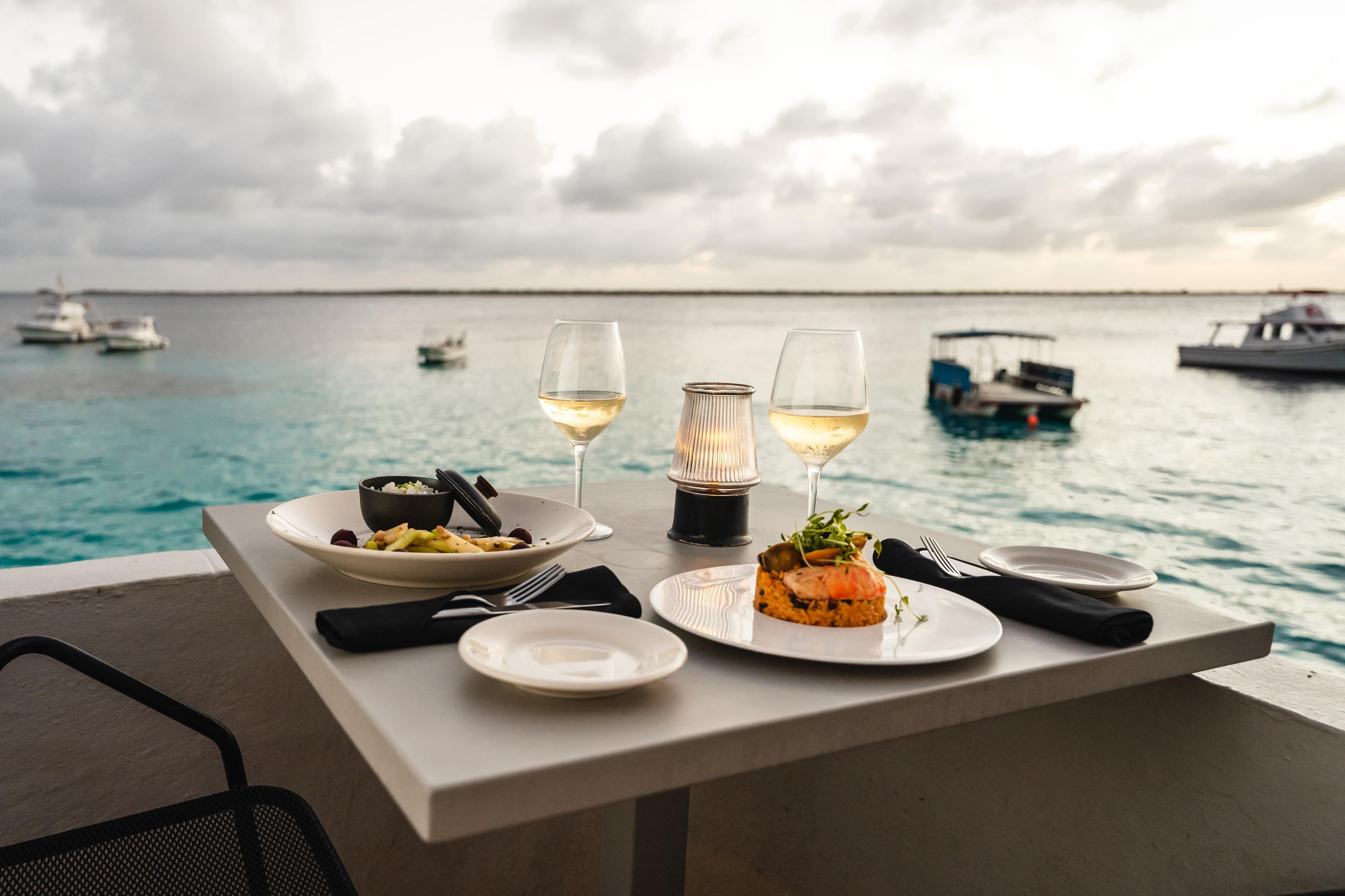 A table set for dining with plates of food, glasses of white wine, and a candle on a balcony overlooking the sea with boats in the background.