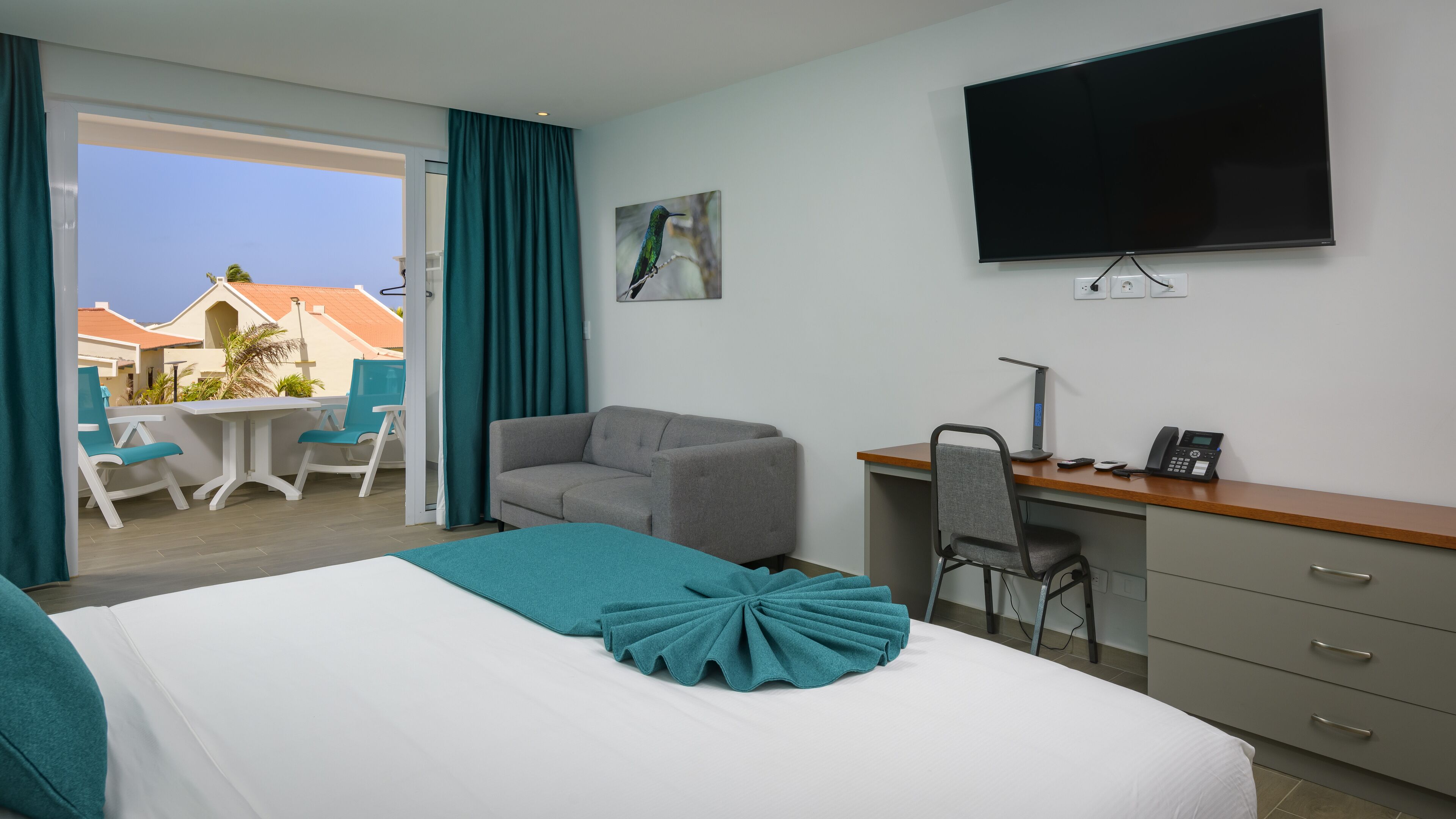 A hotel room with a bed, a desk, a chair, a TV, and a balcony with a view of buildings.