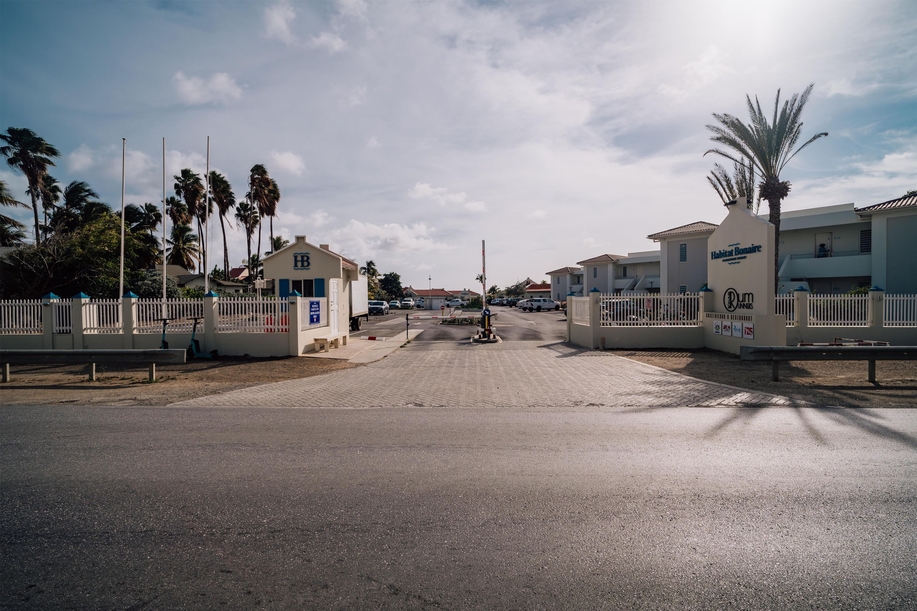 Una entrada cerrada a una zona residencial con palmeras, un camino pavimentado y edificios con señalización.