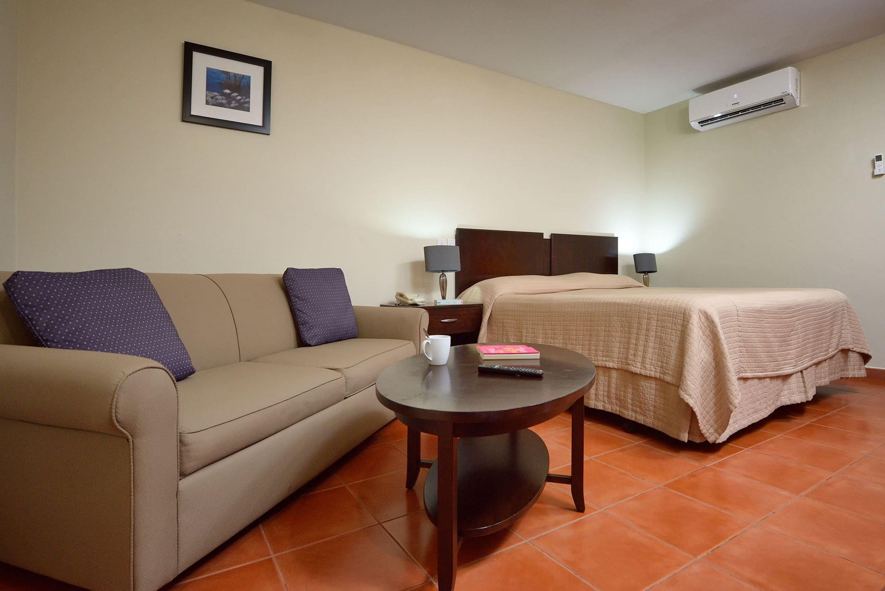 A hotel room with a bed, a sofa, a small table, and an air conditioning unit.