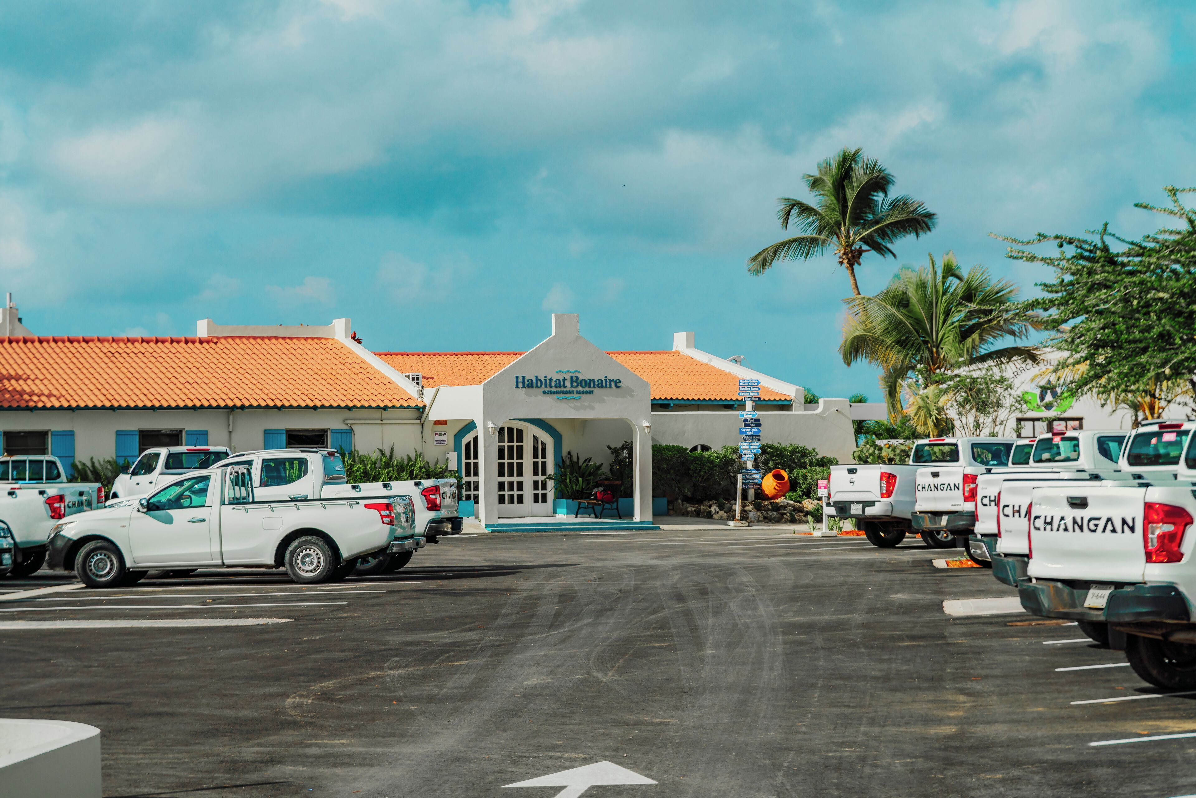 A parking lot with several white pickup trucks parked in front of a building with a red-tiled roof and a sign that reads 'Habitat Boutique.'