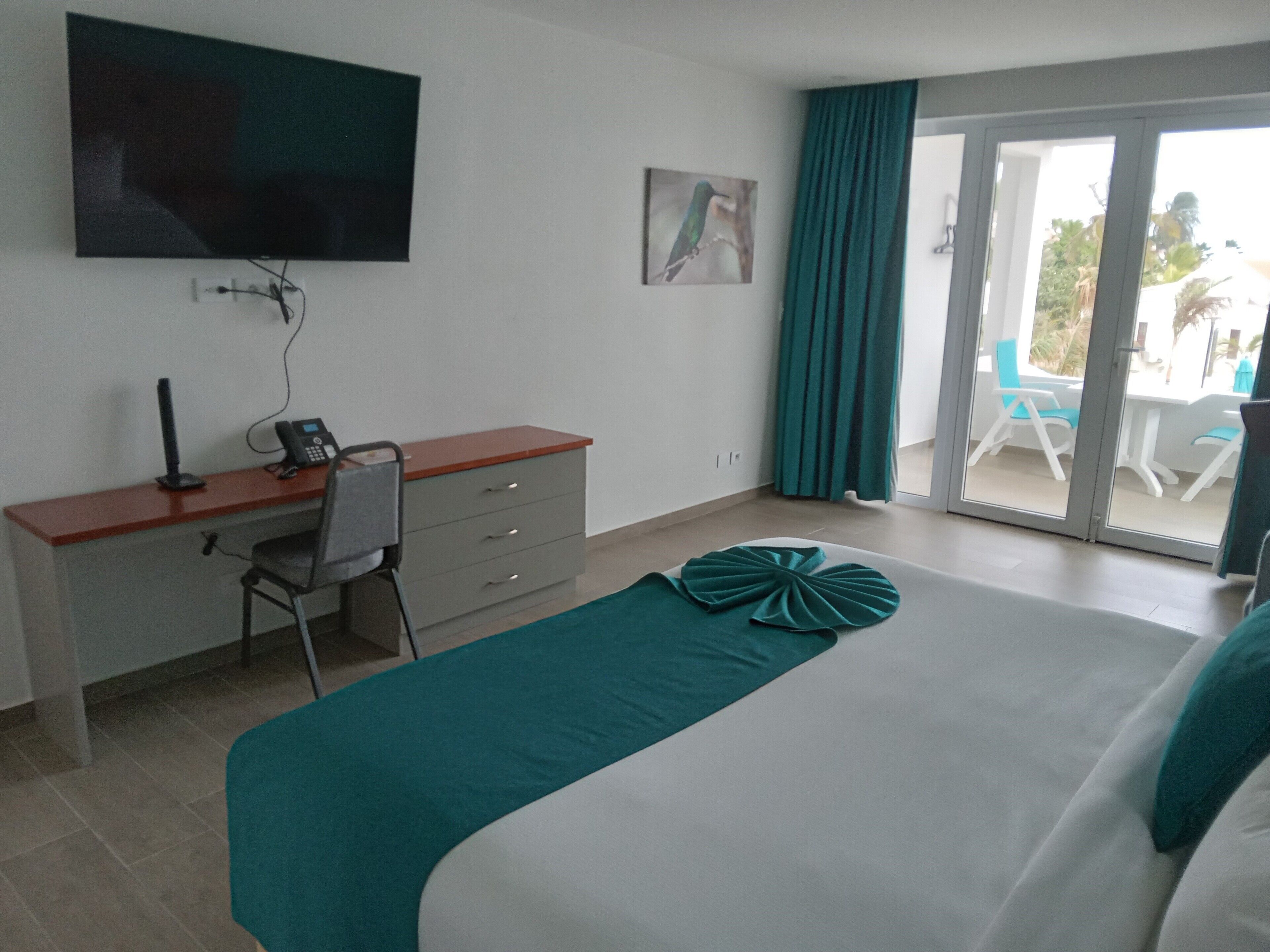 A hotel room with a bed, a desk with a chair, a telephone, a flat-screen TV mounted on the wall, and a sliding glass door leading to an outdoor area with chairs.