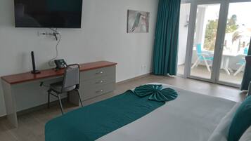Quarto de hotel com uma cama, uma escrivaninha com cadeira, telefone, televisão de tela plana fixada na parede e porta de vidro deslizante que dá para área externa com cadeiras.