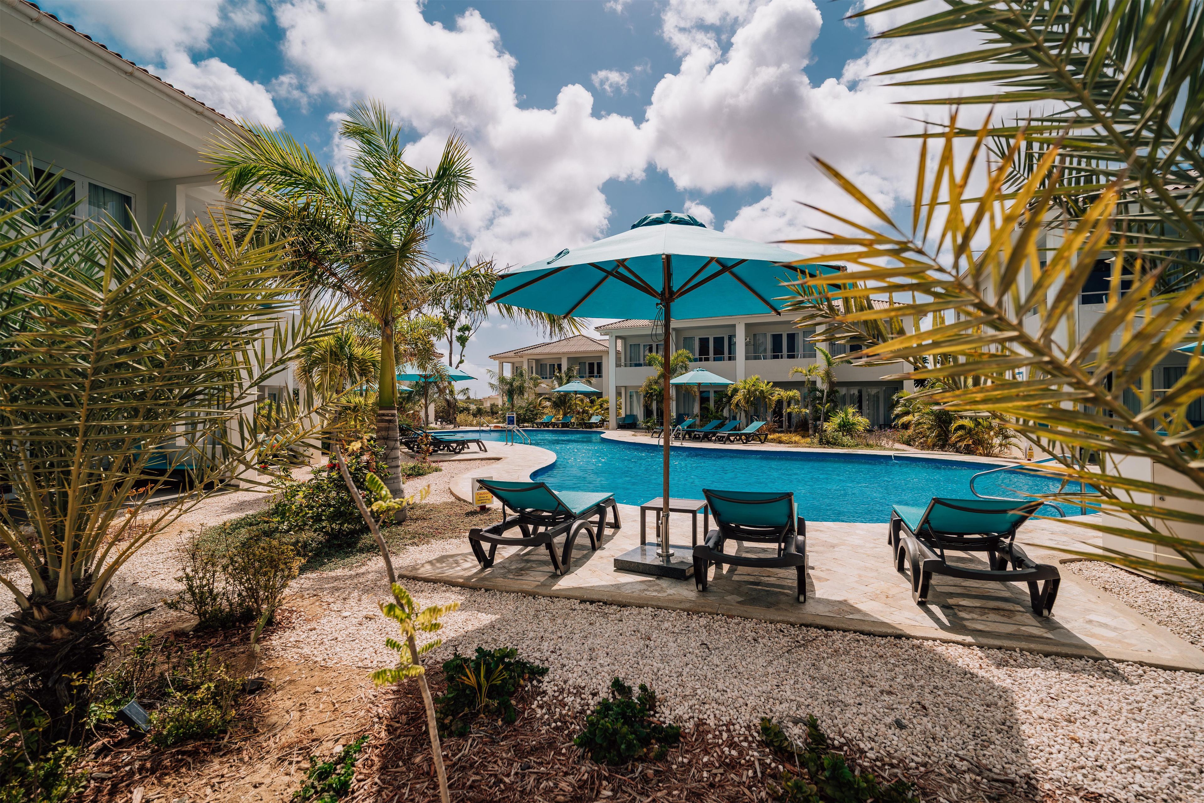 A pool area with lounge chairs and umbrellas, surrounded by palm trees and a building with a tiled roof.