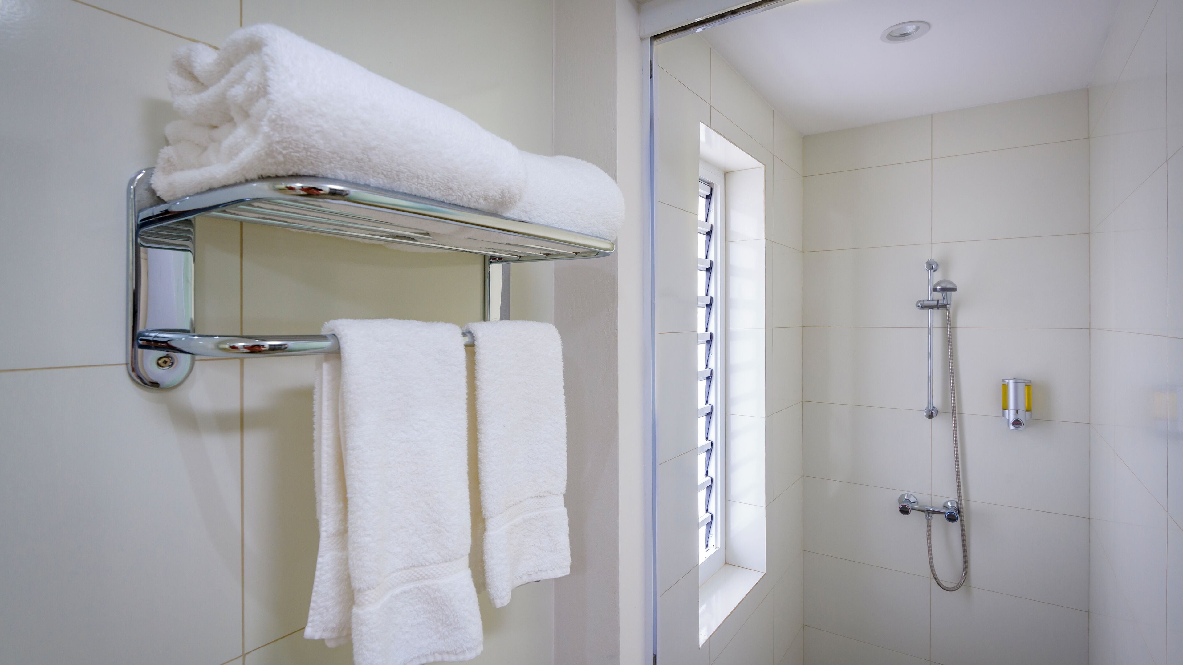 A bathroom with a glass shelf holding a folded white towel, two towels hanging on a rack, and a shower with a handheld showerhead.