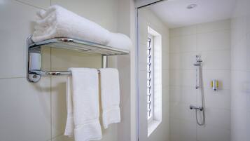 A bathroom with a glass shelf holding a folded white towel, two towels hanging on a rack, and a shower with a handheld showerhead.