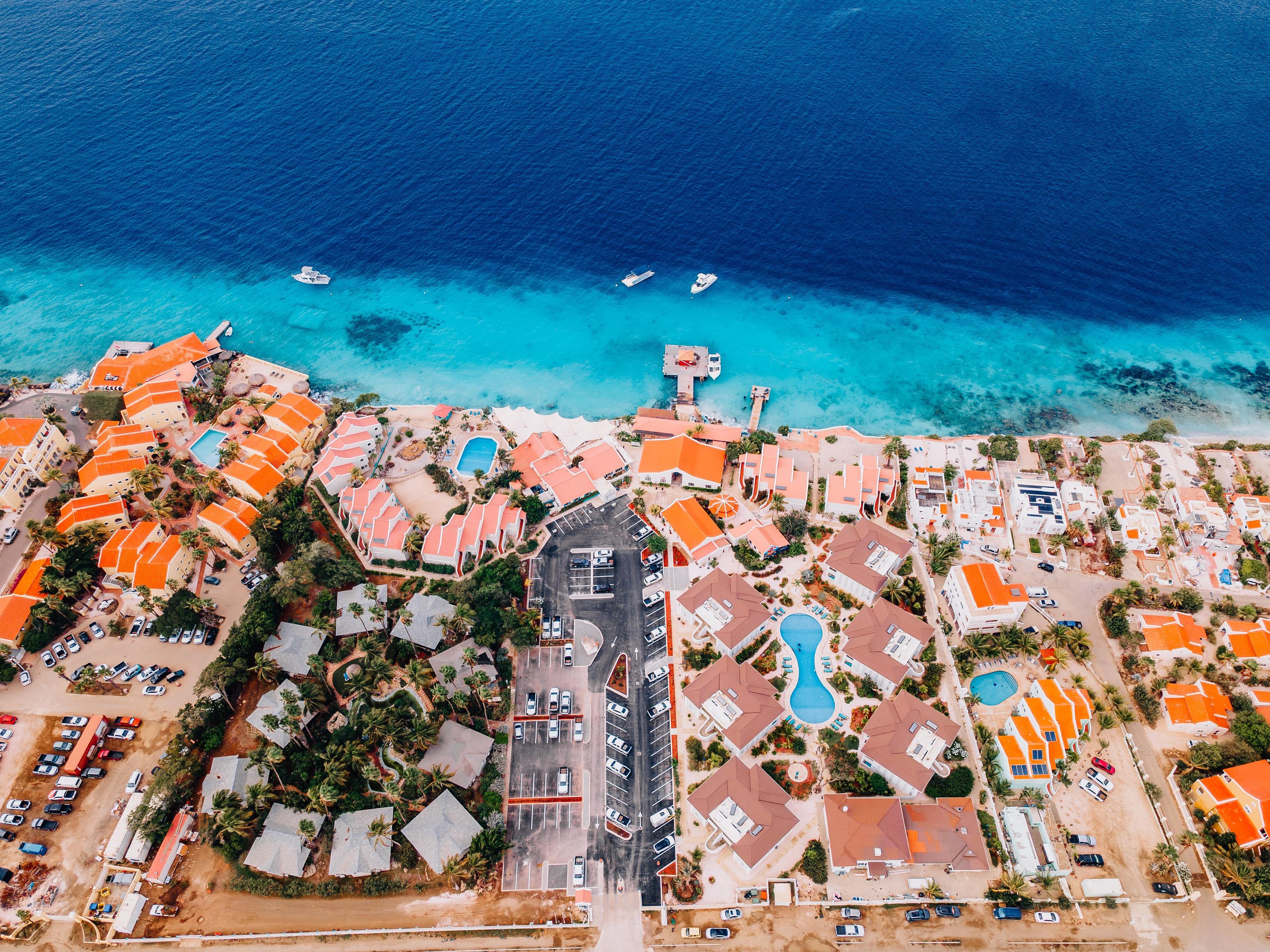 An aerial view of a coastal resort with multiple buildings, a central road, and a clear blue sea.