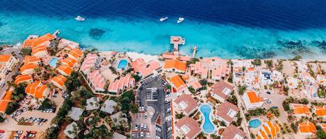 An aerial view of a coastal resort with multiple buildings, a central road, and a clear blue sea.