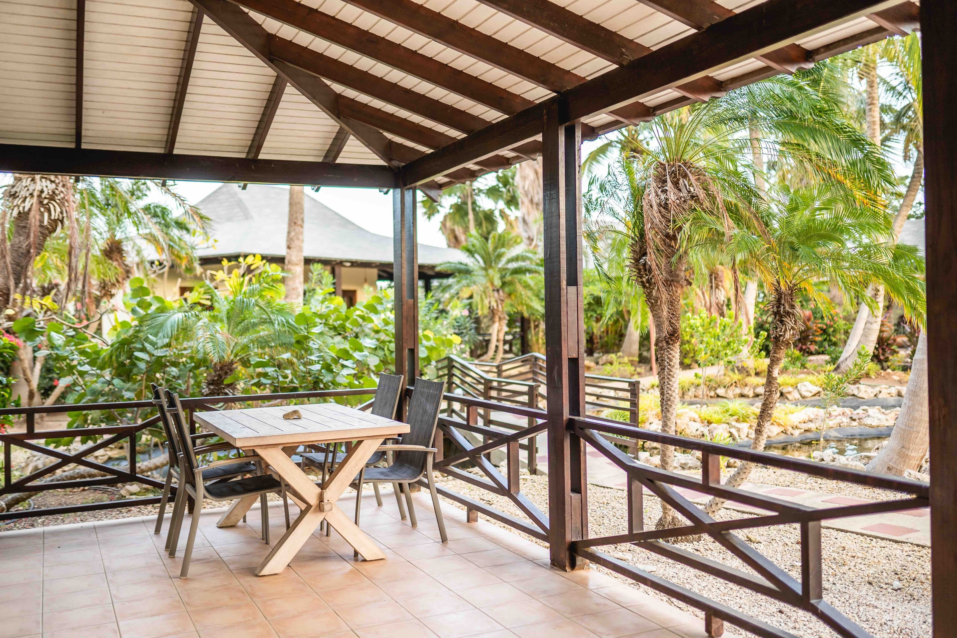 Un espace extérieur aménagé avec une zone salon couverte, comprenant une table et des chaises en bois, le tout entouré de palmiers et de verdure.