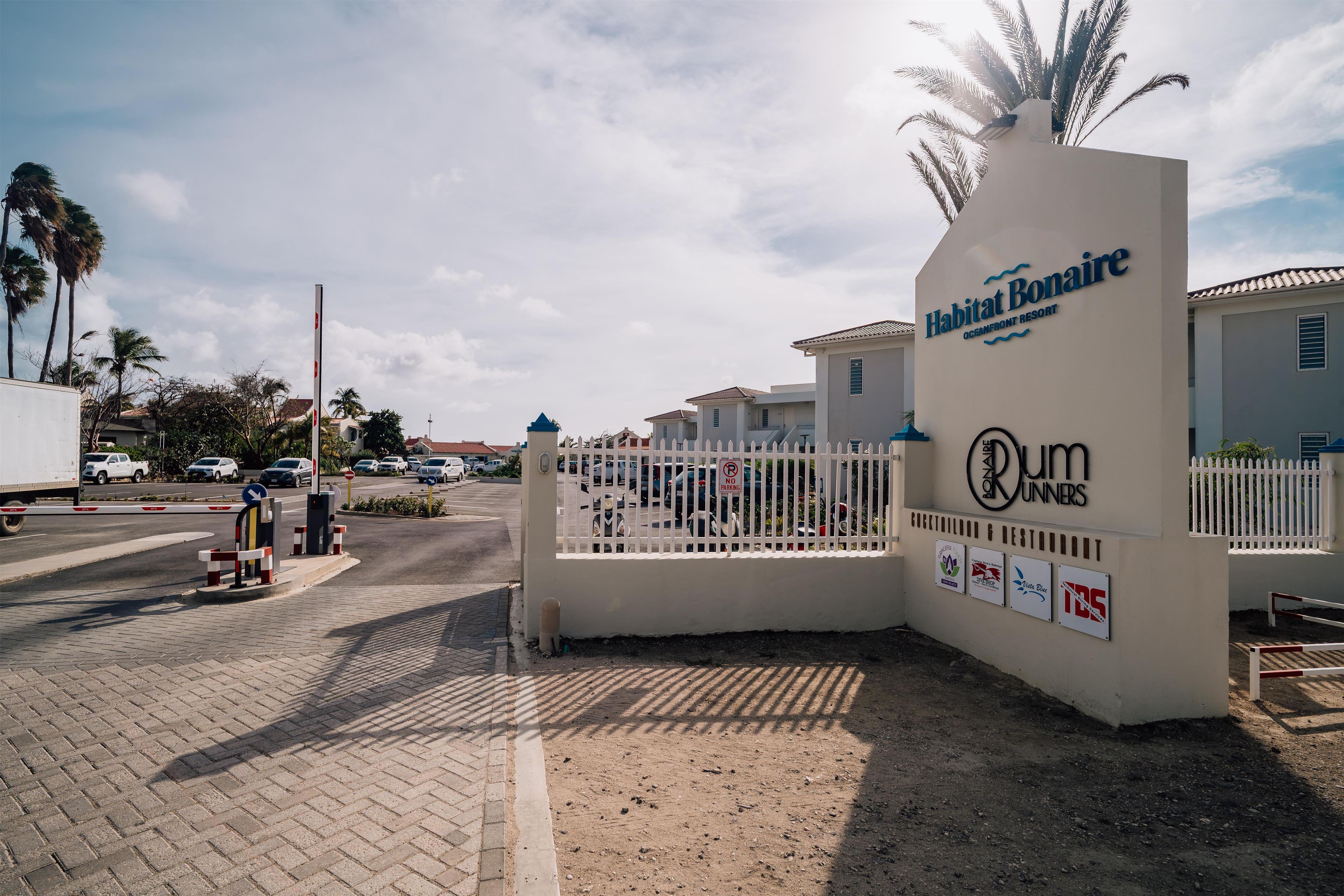 Une entrée fermée par un portail mène à une propriété où l’on peut voir une enseigne indiquant « Habitat Bonaire » et le restaurant « Rum Runners ».