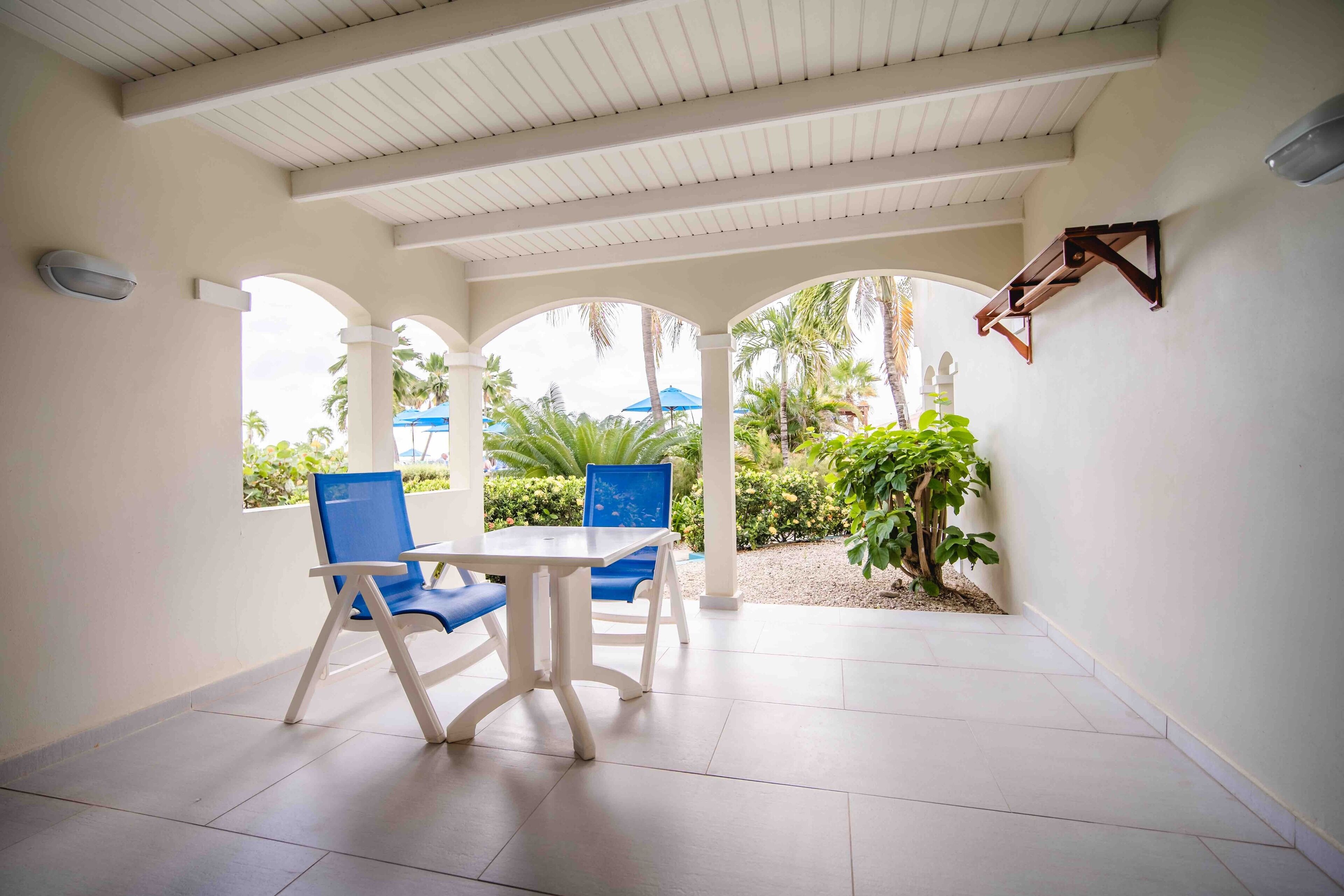 A covered patio area with a white ceiling, white walls, and a tiled floor. There are two blue chairs and a white table.