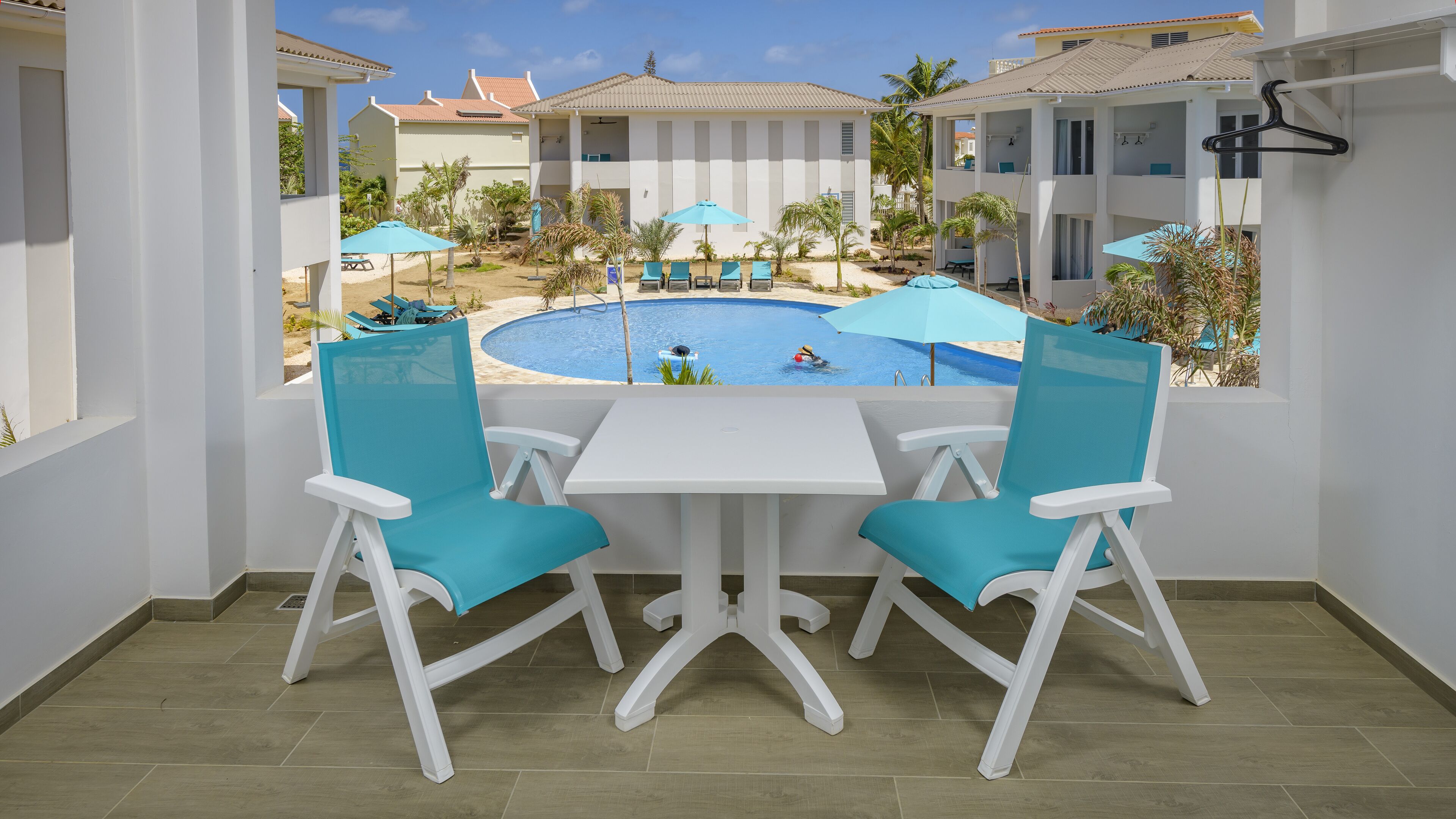 A balcony with a view of a swimming pool, white chairs, and a table.