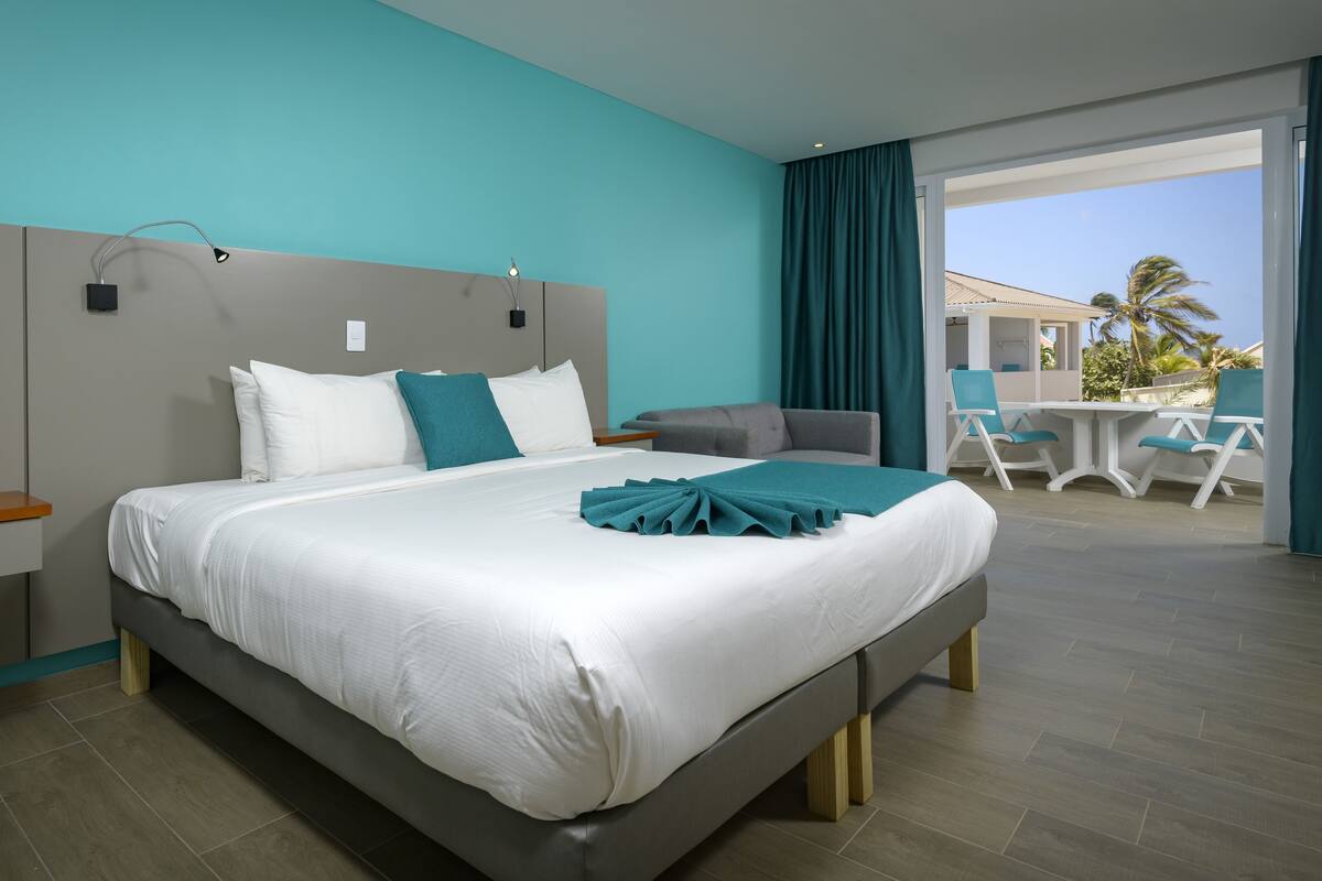 A modern hotel room with a large bed, a desk, and a balcony with outdoor seating.
