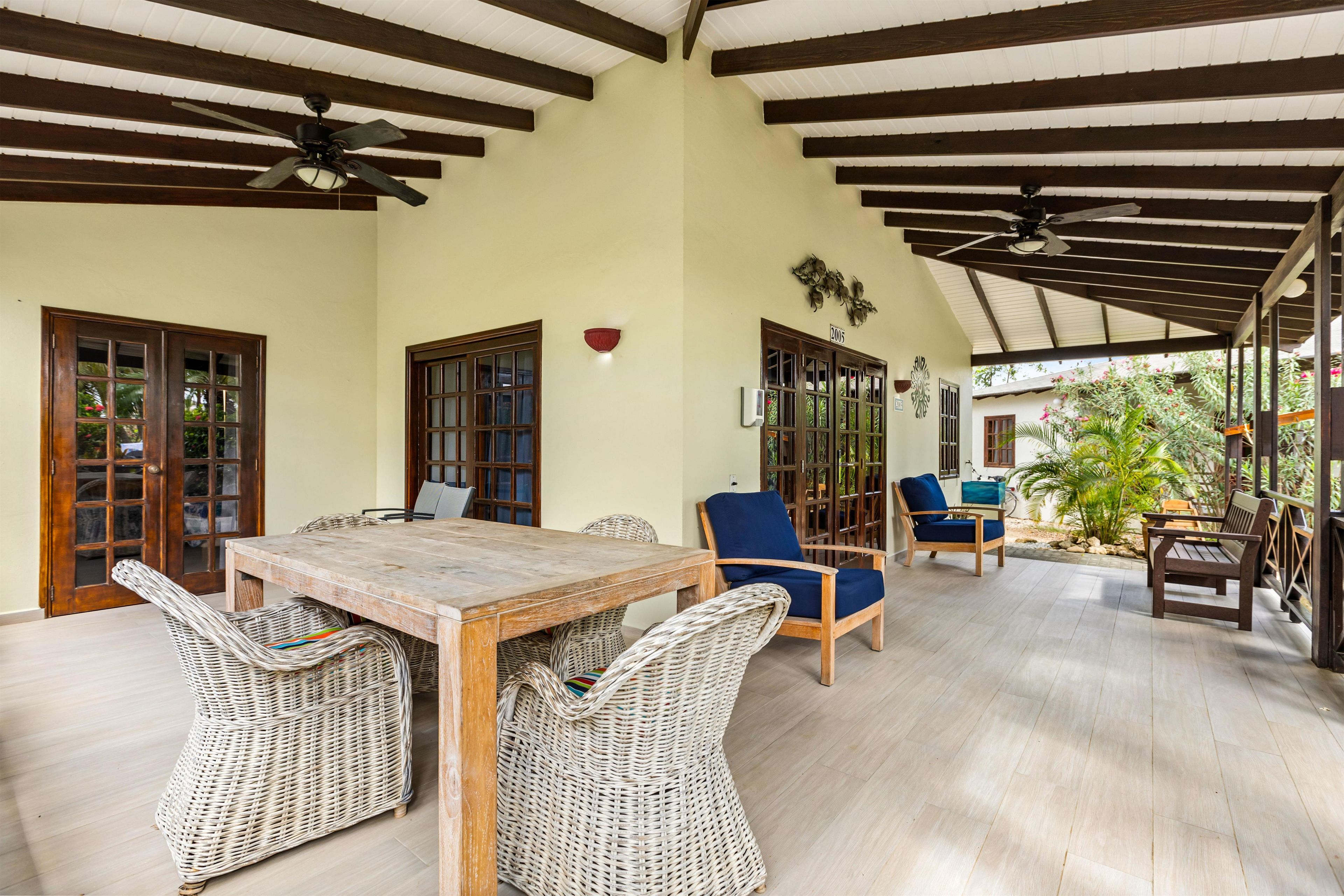 Un espacio al aire libre techado con una mesa de madera y sillas de mimbre, un ventilador de techo y vistas a un jardín.