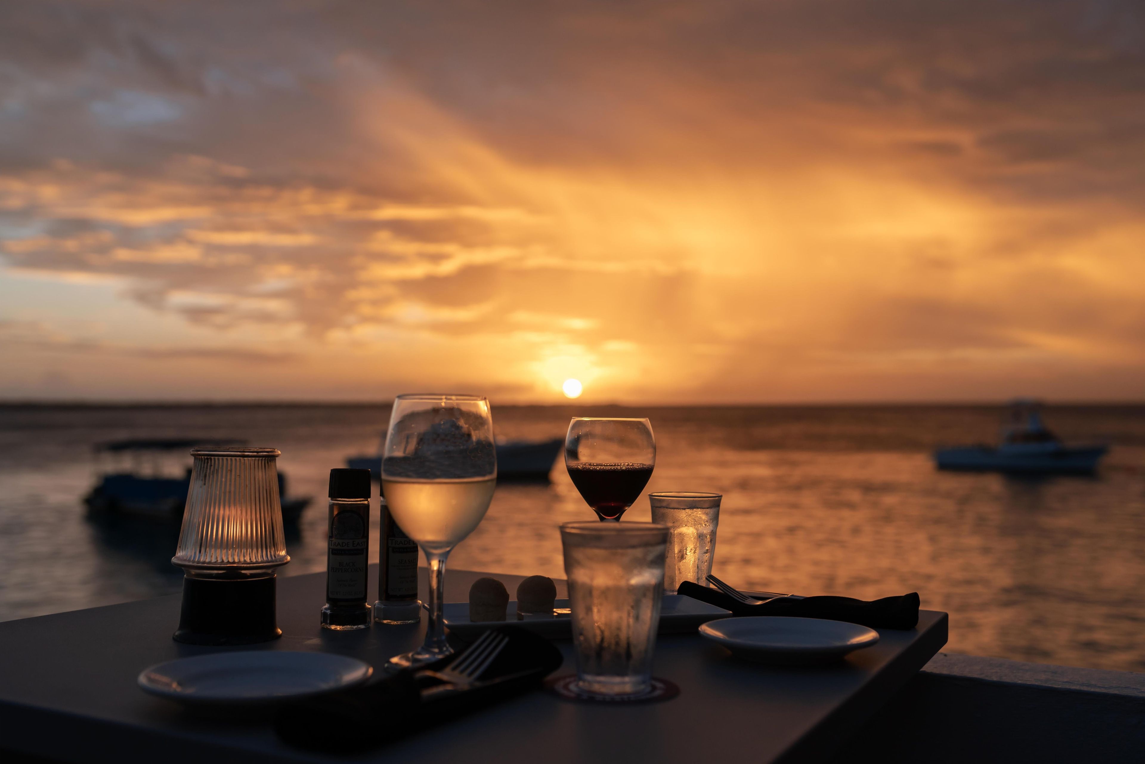 A table set with glasses, a bottle, and cutlery by the sea at sunset.