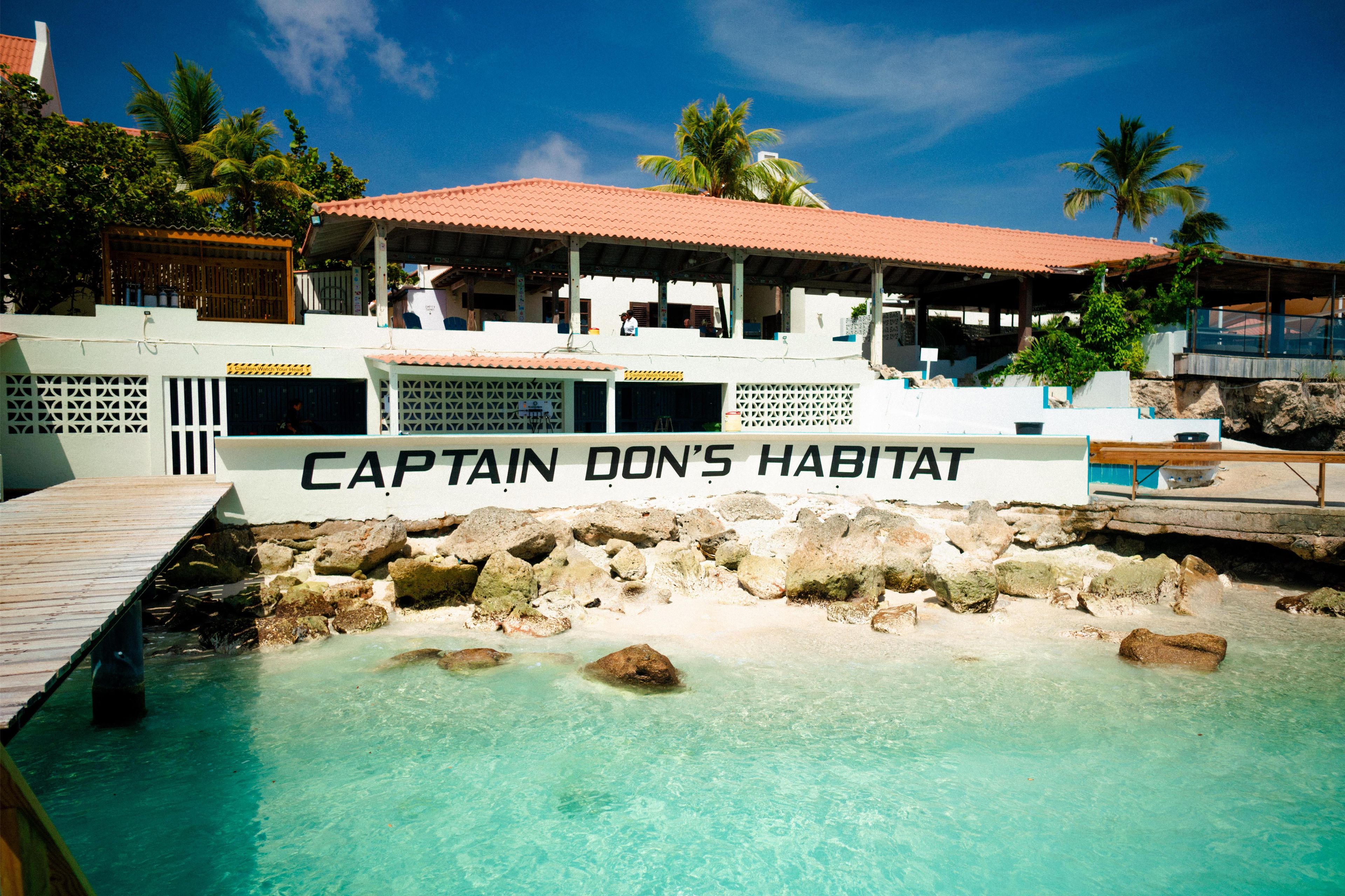 Een resort met een zwembadgedeelte genaamd 'Captain Don's Habitat', met een helder zwembad omringd door rotsen en een houten terras.