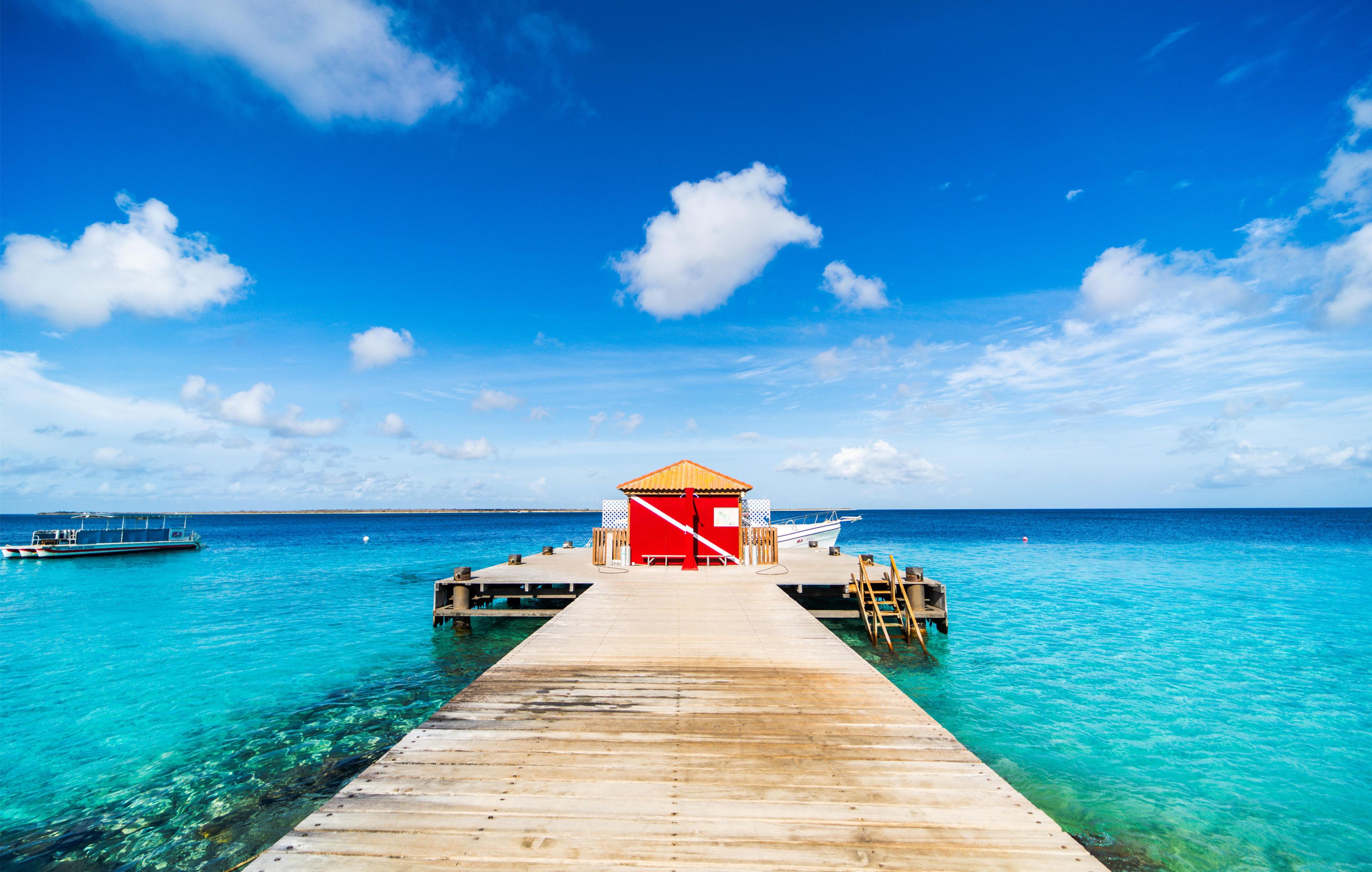 A wooden pier leading to a red and white building with a thatched roof, extending into a turquoise sea.