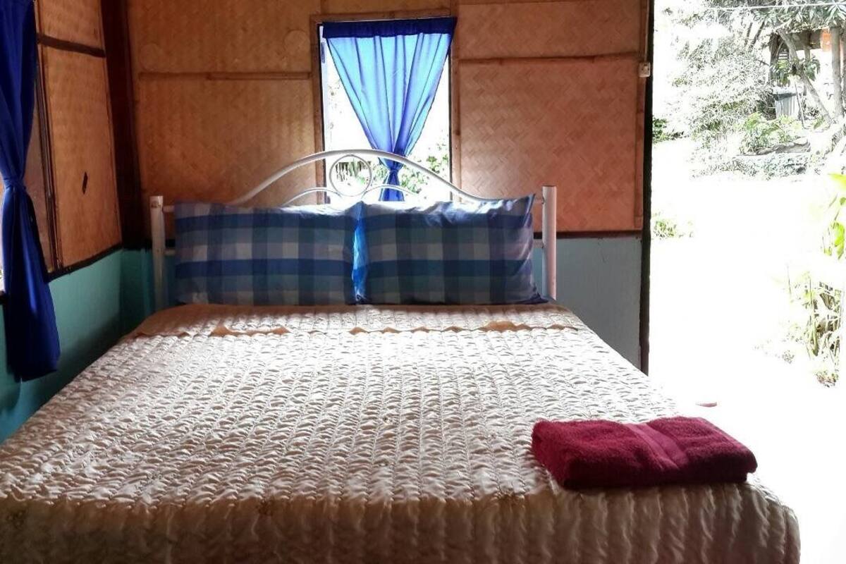 A bed with a checkered pillow and a red towel, a window with blue curtains, and a view of greenery outside.