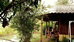 A rustic wooden structure with a thatched roof, situated beside a calm pond, surrounded by lush greenery. - Rainbow House (Pai)