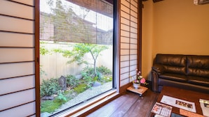 A traditional Japanese room with tatami flooring, a black leather sofa, a sliding glass door leading to a garden, and a small table with a vase of flowers.