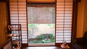 A traditional Japanese room with tatami flooring, a low wooden desk, a bookshelf, and a view through shoji screens to a garden.