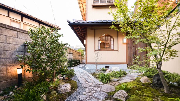 A traditional Japanese-style garden with a stone pathway leading to a building with a tiled roof and wooden gates.