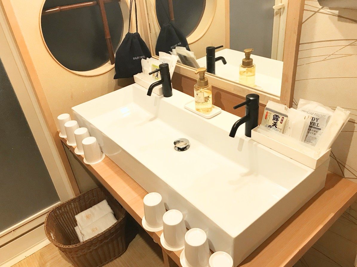 A bathroom with a white sink, two black faucets, a mirror, and a wooden shelf with white cups.