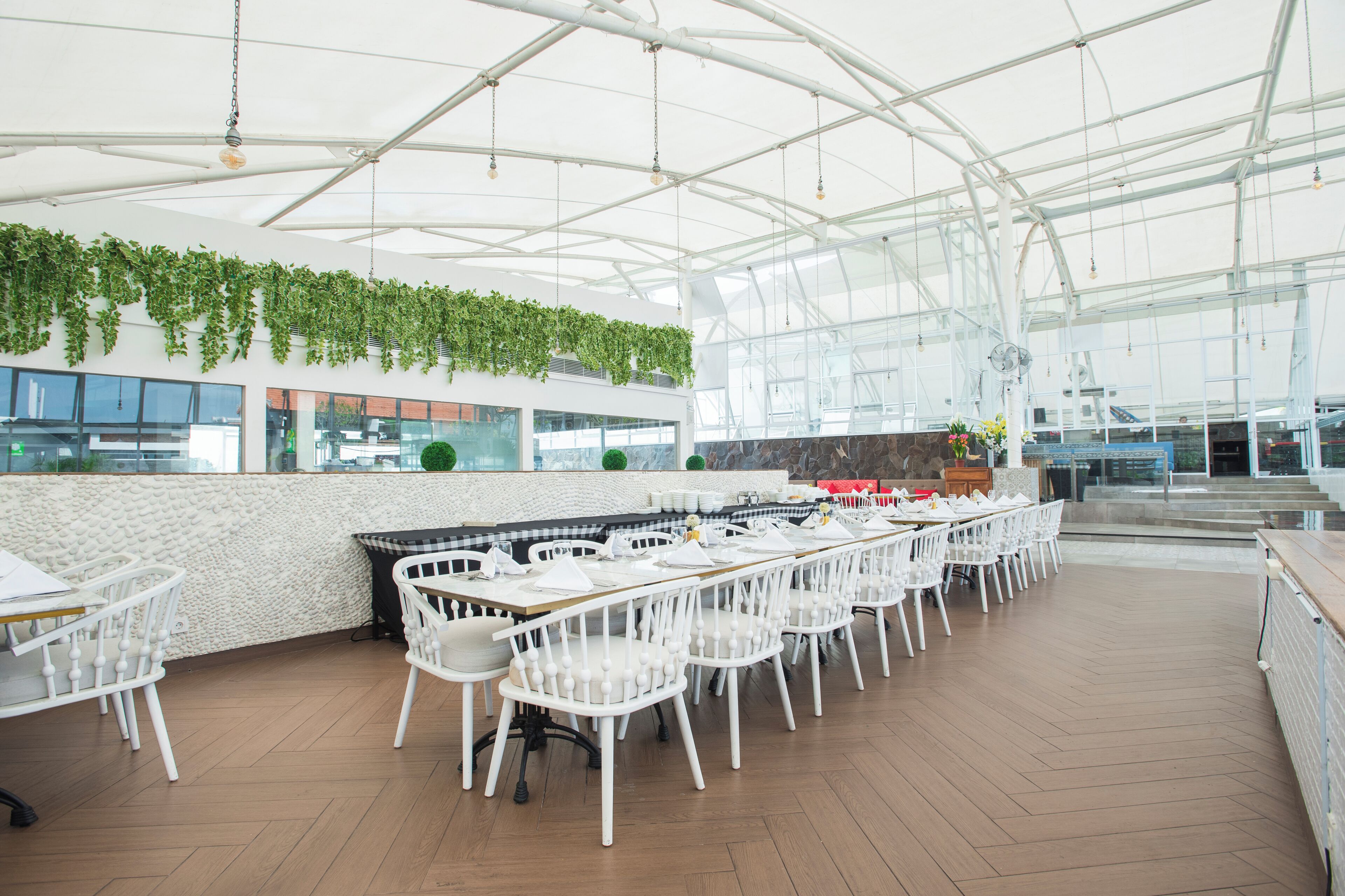 Un amplio comedor interior con sillas y mesas blancas, un techo alto con luces colgantes y vistas al exterior a través de grandes ventanales.