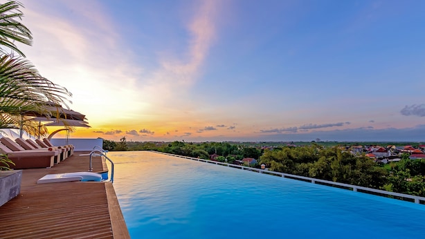 Une piscine entourée de chaises longues et d’une terrasse en bois, le tout bordé de verdure et avec des maisons en arrière-plan.