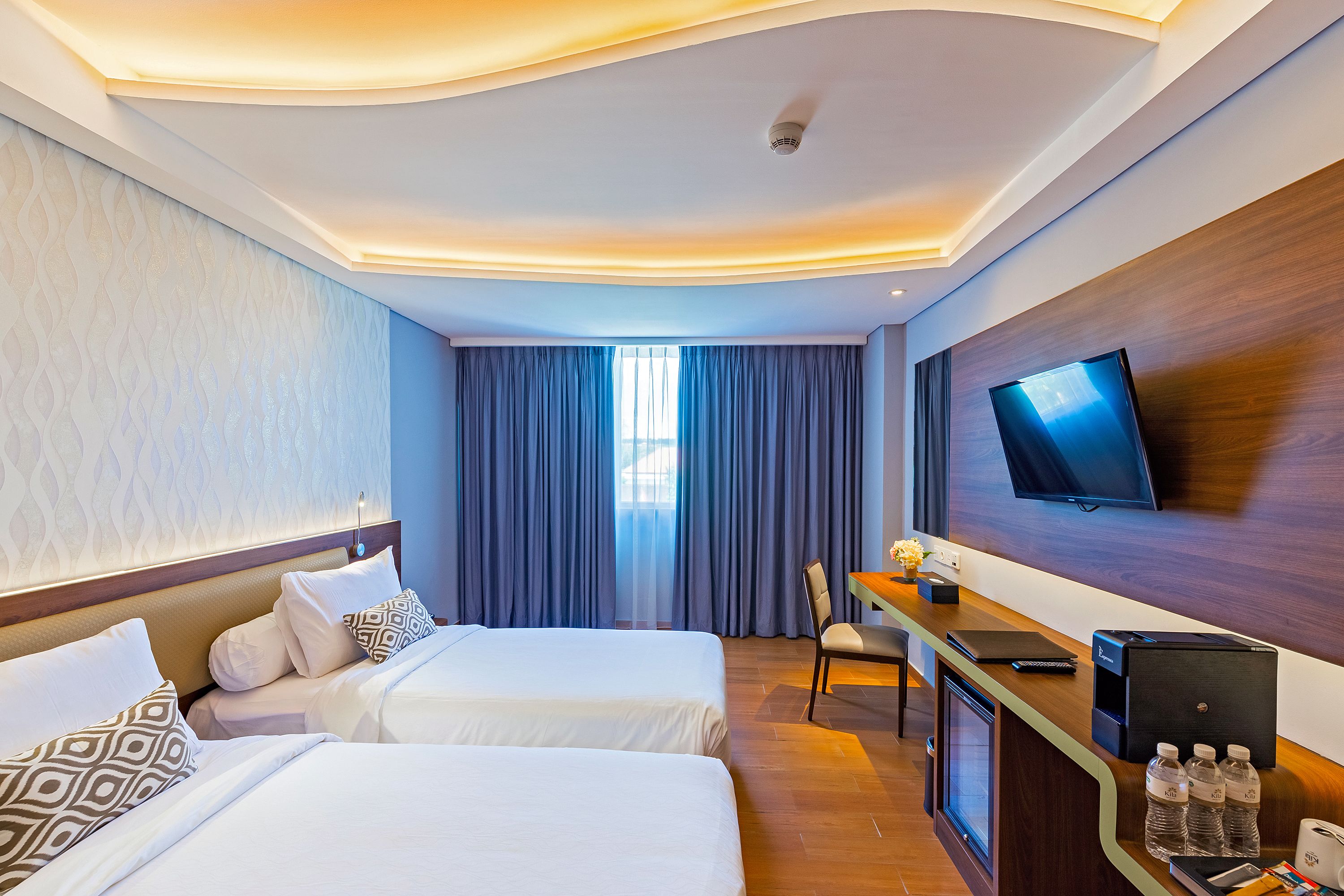 A hotel room with two beds, a desk with a computer, a flat-screen TV, and a minibar.